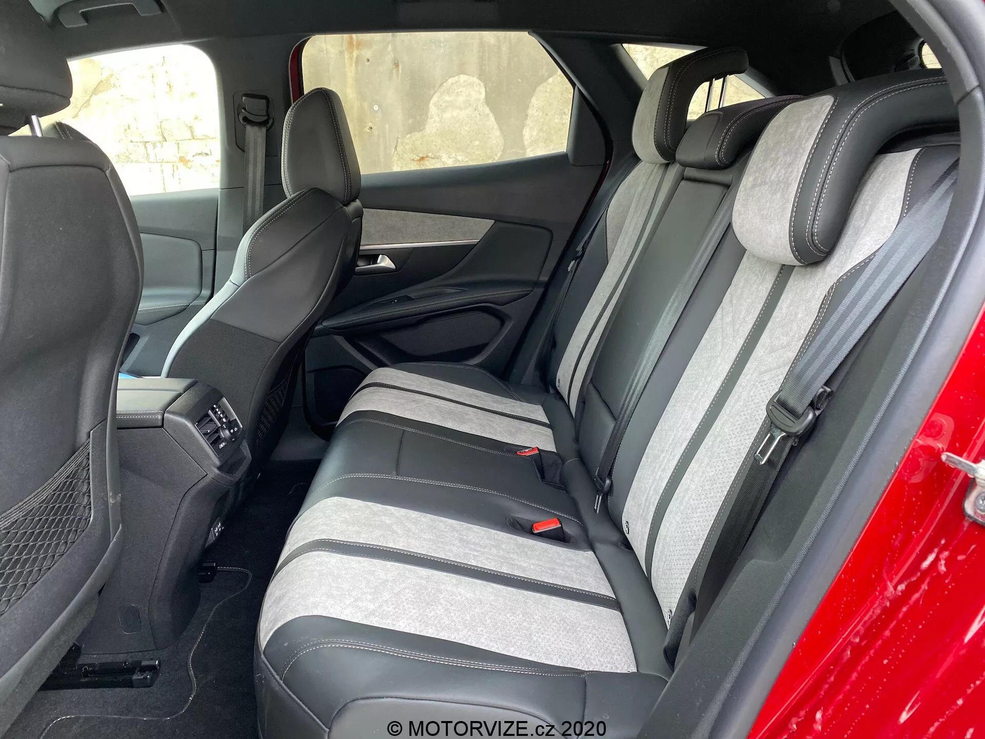 Photo de l'intérieur d'une Peugeot 3008 2021 se concentrant sur la zone des sièges arrière, vue de l'extérieur et en angle vers le côté conducteur. Les sièges arrière sont en tissu bicolore gris foncé et clair avec un motif à rayures texturées. Les appuie-têtes correspondants sont visibles. Une partie des sièges avant et de la console centrale est également visible. Les sièges arrière ont des ceintures de sécurité à trois points et un accoudoir avec porte-gobelets intégrés. Les panneaux de porte correspondent aux couleurs des sièges avec des grilles de haut-parleurs et des sections de contrôle de puissance.