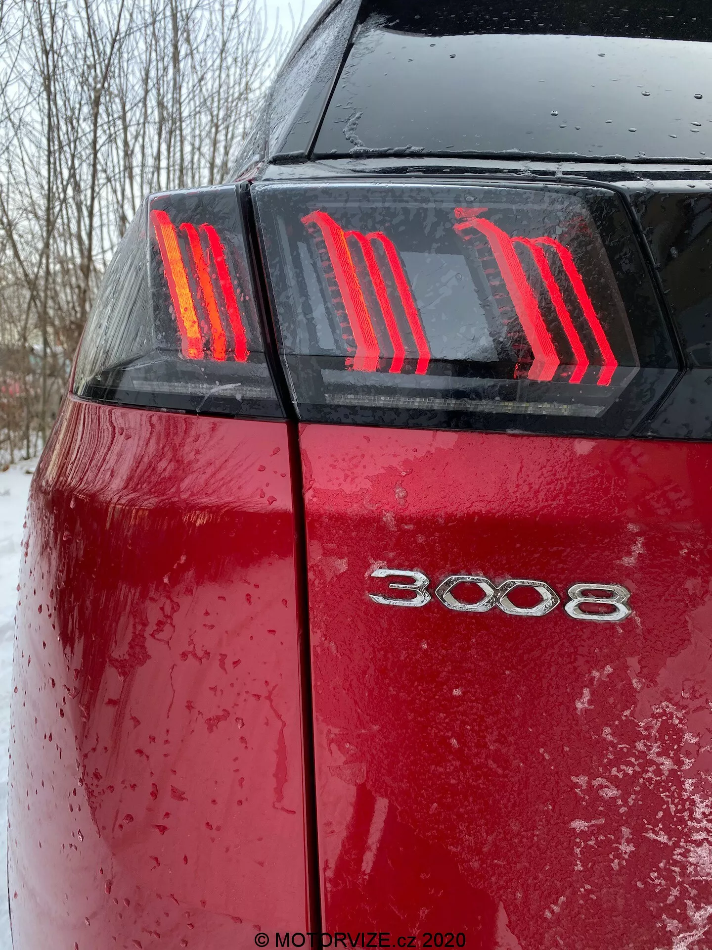Gros plan sur la partie arrière d'une Peugeot 3008 2021 rouge vue sous un angle bas, mettant en valeur les feux arrière distinctifs à trois bandes verticales en forme de griffes et le badge du modèle '3008'. La surface mouillée de la voiture, couverte de gouttes d'eau et de saleté ou de neige, met en évidence une utilisation récente par mauvais temps avec des conditions lumineuses nuageuses.