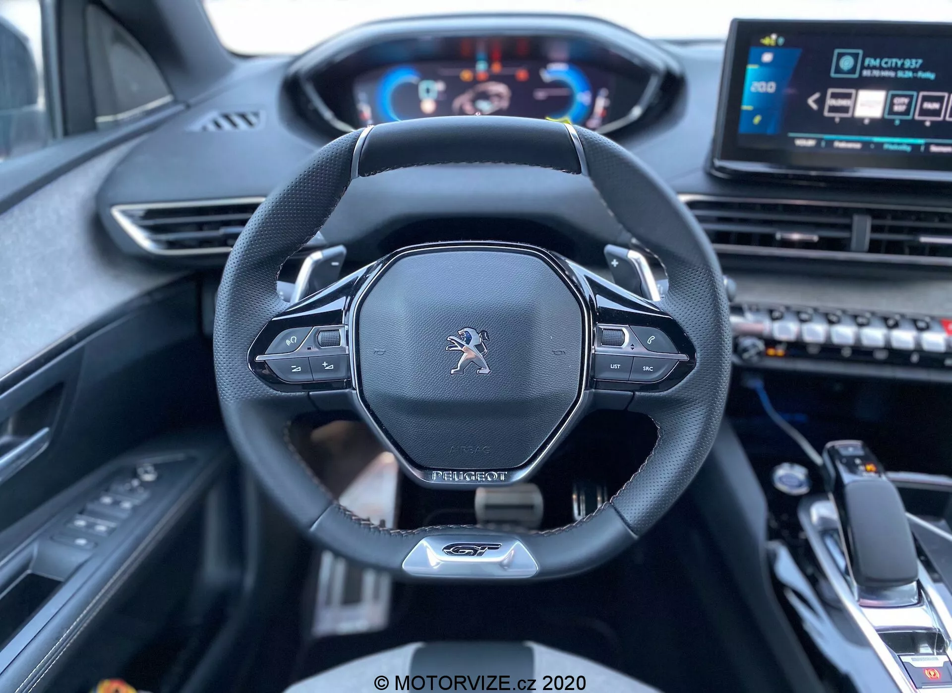 Vue intérieure depuis le siège conducteur d'une Peugeot 3008 2021, montrant le volant et le tableau de bord. L'image se concentre sur le volant encadré au centre, arborant le logo Peugeot, un design moderne avec une partie inférieure plate, un bord en cuir et des commandes intégrées probablement pour les réglages audio et du régulateur de vitesse. Derrière le volant, des palettes pour le changement de vitesse manuel sont visibles. Un tableau de bord numérique affiche le compteur de vitesse et le compte-tours, illuminé pour indiquer que le véhicule est allumé. À droite du volant, un système d'info-divertissement à écran tactile et divers boutons de contrôle sont partiellement visibles, ainsi qu'un sélecteur de vitesse et un accoudoir central au premier plan inférieur, suggérant un véhicule moderne et technologiquement avancé avec un intérieur haut de gamme.