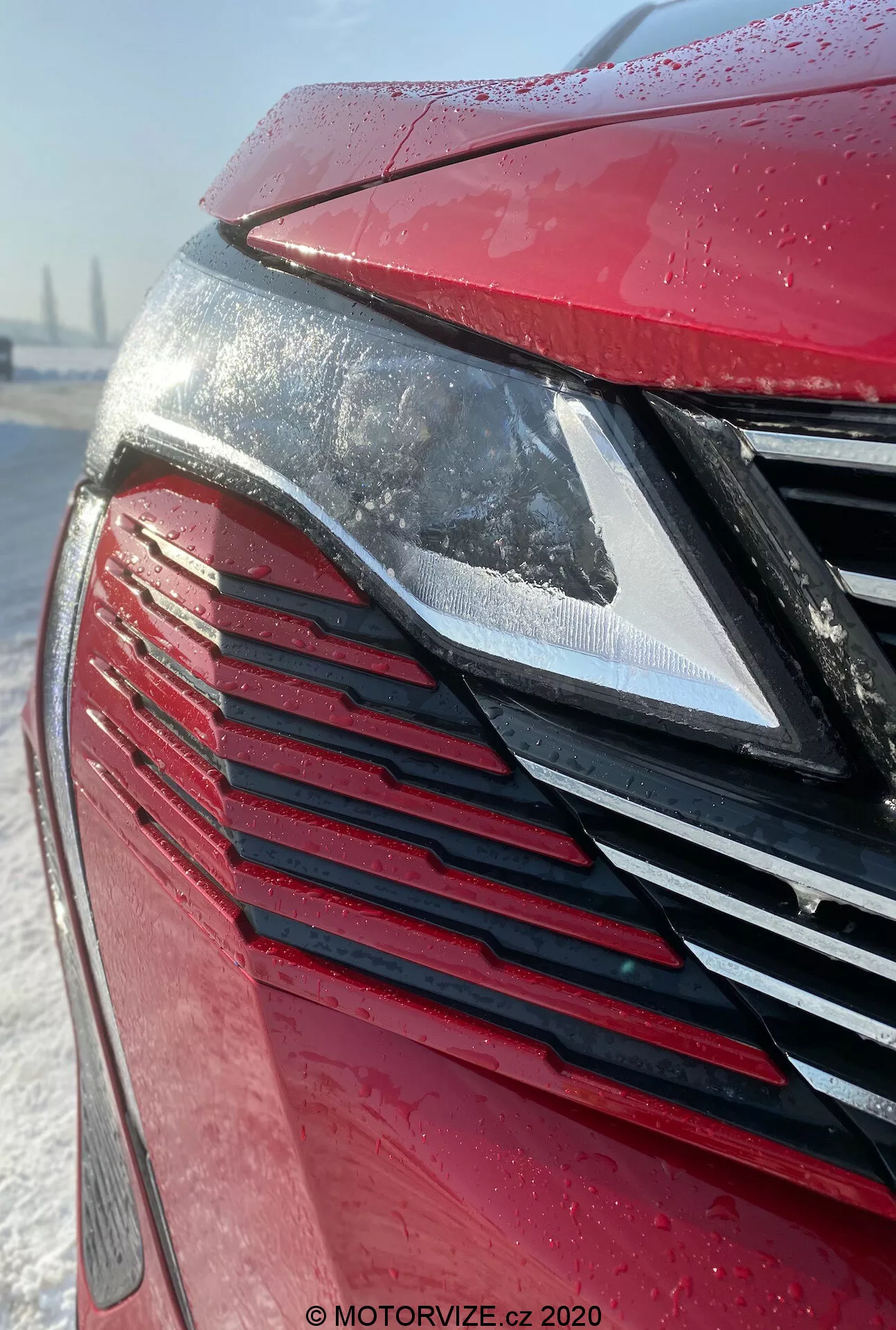 Vue rapprochée de l'avant de la Peugeot 3008 2021, mettant en valeur le phare LED moderne et la calandre avec des lamelles verticales dans une finition rouge métallisée couverte de gouttelettes d'eau, suggérant une pluie récente ou du givre. L'arrière-plan enneigé suggère des conditions hivernales ou un entretien soigné du véhicule.