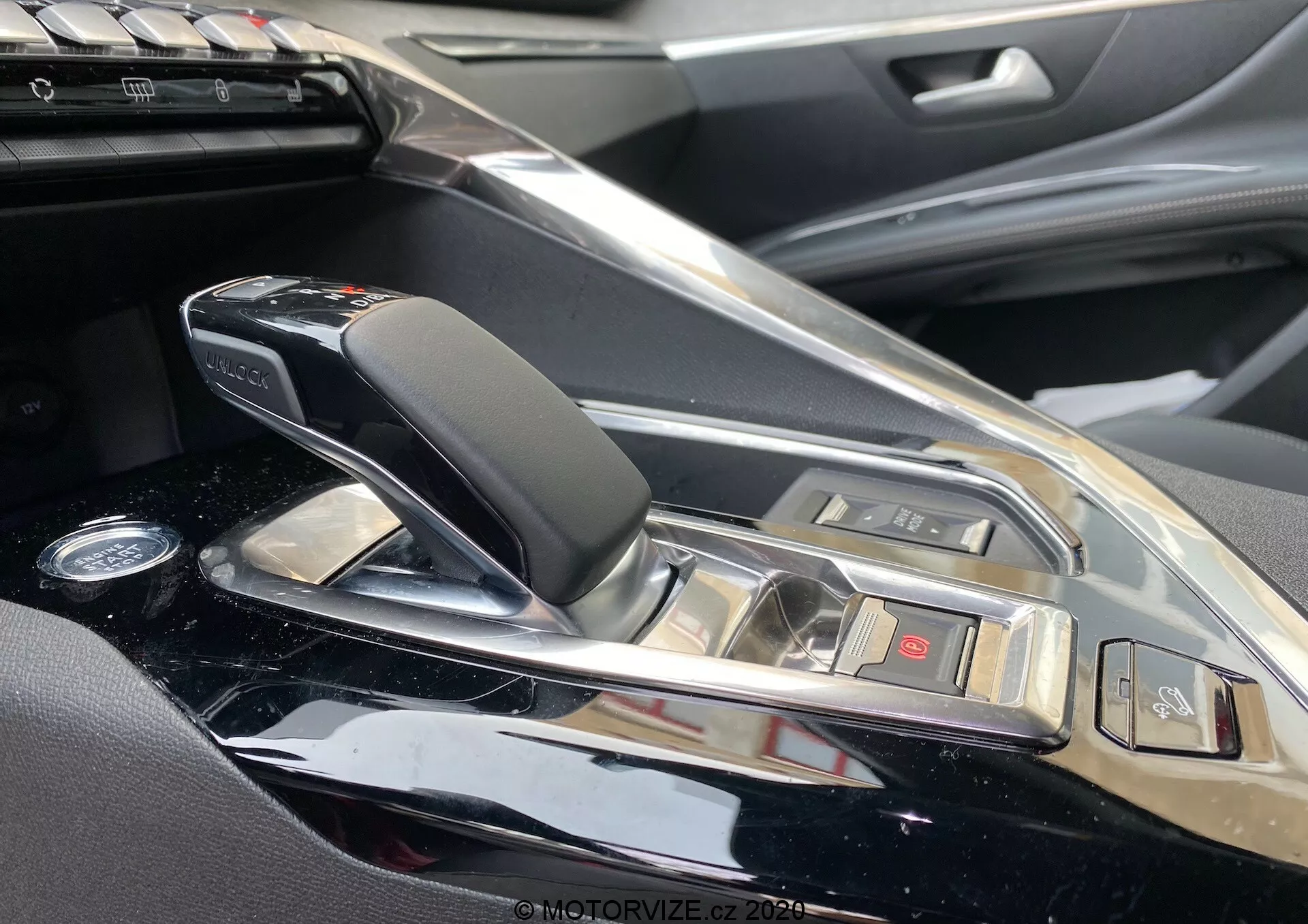 Vue en gros plan de la console centrale et de la zone du levier de vitesses de la Peugeot 3008 2021 depuis le côté passager. Il comporte un levier de vitesses automatique moderne et élégant avec des accents en cuir et en chrome, entouré d'une finition brillante. Des boutons pour diverses fonctions du véhicule et un aperçu du système d'infodivertissement sont visibles, indiquant un intérieur de voiture haut de gamme avec des matériaux de haute qualité et des caractéristiques technologiques sophistiquées.