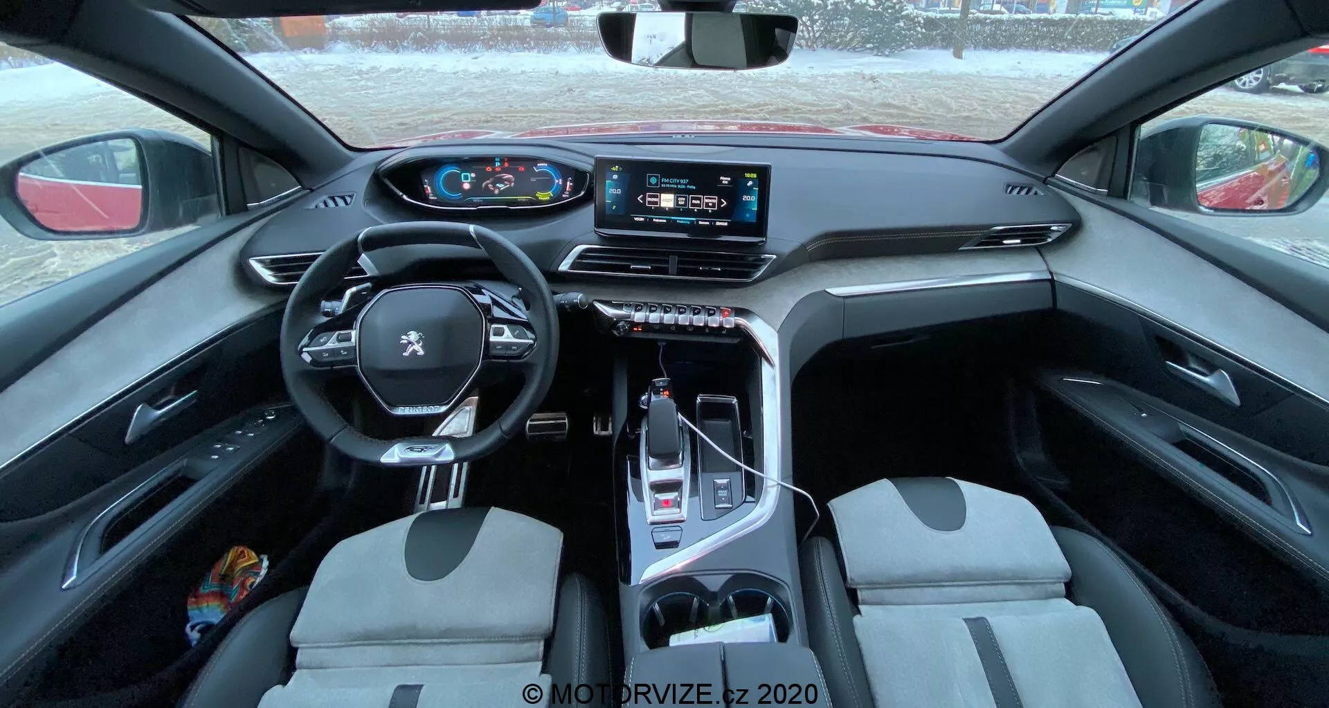 Vue intérieure de la Peugeot 3008 2021, mettant en avant un volant multifonction en cuir avec commandes intégrées, un tableau de bord numérique, un grand écran tactile d'infodivertissement sur la console centrale, un sélecteur de vitesses électronique, des sièges avant bicolores et un mélange de matériaux pratiques et luxueux. La photo est prise depuis le siège arrière, offrant un aperçu expansif de l'habitacle, soulignant le design et les caractéristiques technologiques.