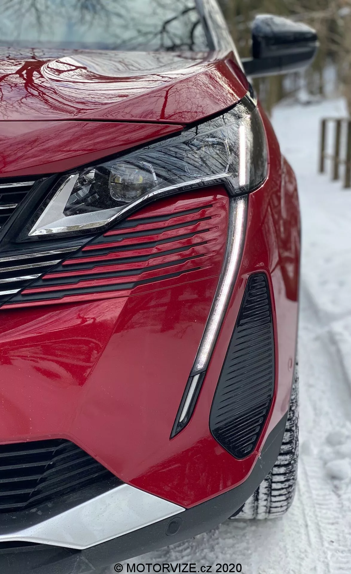 Vue en gros plan en contre-plongée d'une Peugeot 3008 2021 rouge moderne. L'accent est mis sur le quart avant-gauche mettant en valeur des phares LED angulaires élégants, une grande calandre agressive, un design de ventilation marqué, une finition peinture brillante et des panneaux de carrosserie aux contours acérés. Le rétroviseur latéral intègre des clignotants et la scène inclut un sol enneigé, suggérant des conditions hivernales. Filigrane de MOTORVIZE.cz visible.