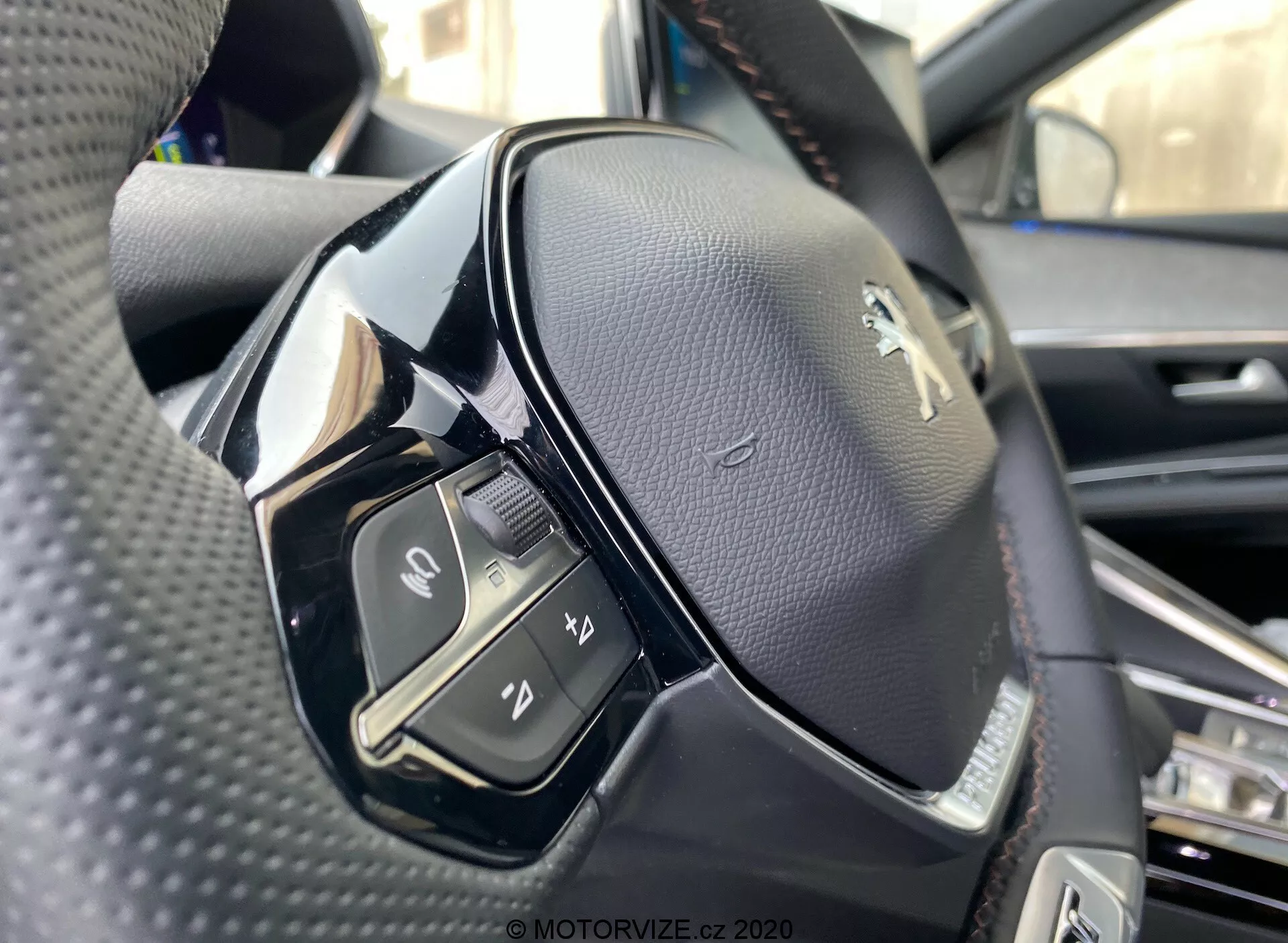 Vue en gros plan d'un volant de la Peugeot 3008 2021 depuis la perspective du siège conducteur. L'image accentue le côté gauche du volant, prise d'un angle légèrement bas et capturant les détails des commandes du côté gauche et le logo Peugeot. Le volant est recouvert de cuir noir perforé avec un mélange de matériaux noirs mats et brillants. Les commandes visibles comprennent un interrupteur, des boutons pour les fonctions multimédia et de communication comme accepter ou terminer des appels, l'activation de la commande vocale et une autre fonction non identifiée. La mise au point est nette, montrant les coutures du cuir et la finition brillante sur certaines parties en plastique, éventuellement des boutons tactiles.