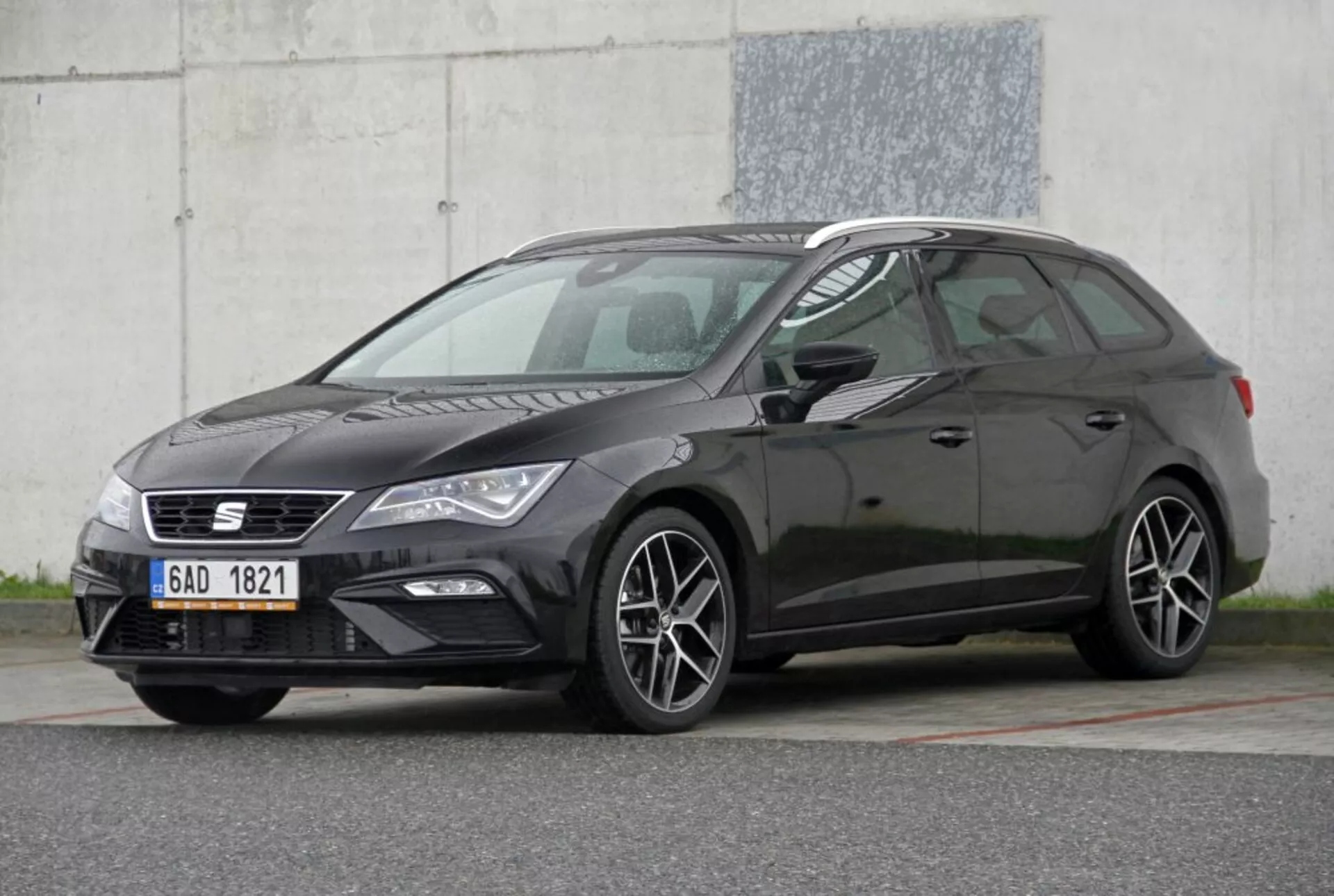 Véhicule hatchback Peugeot 308 SW de couleur sombre de 2013 mettant en avant ses caractéristiques : grille étroite avec emblème, unités de phares élégants avec feux de jour, jantes en alliage à plusieurs branches suggérant un modèle ou un niveau de finition sportif, profil latéral distinctif de hatchback avec la ligne de toit descendante, dans une couleur de carrosserie sombre contrastante, pris sous un angle frontal de trois quarts contre un fond industriel