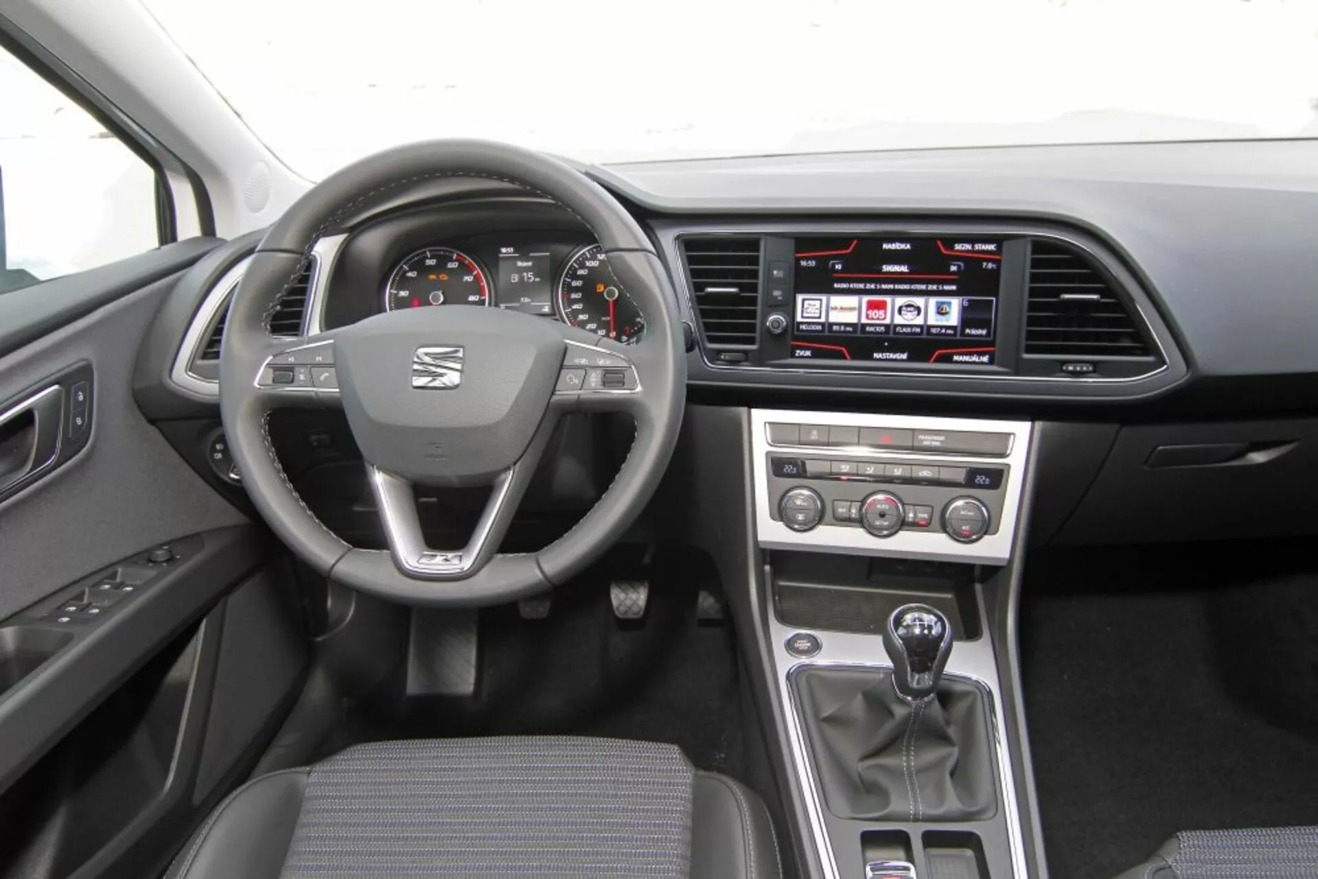 Vue intérieure d'une Peugeot 308 SW de 2013 montrant le volant avec logo et commandes multifonctions, compteur de vitesse et compte-tours analogiques avec affichage numérique, système d'infodivertissement tactile sur le tableau de bord, boutons de contrôle manuel du climat sur la console centrale, levier de vitesse manuel et garniture en tissu foncé pratique.