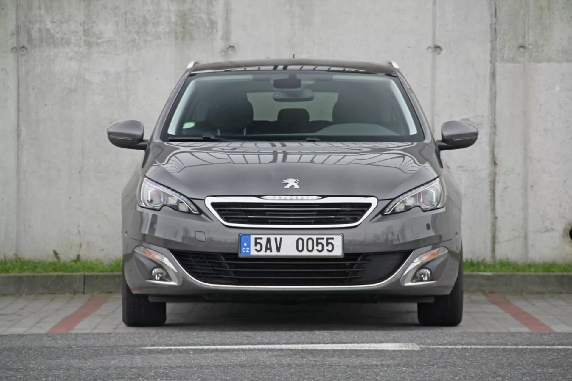 Vue frontale d'une Peugeot 308 SW de 2013 en gris foncé avec grille chromée, phares élégants, feux de brouillard, pare-chocs avant sportif, capot avec plis doux, essuie-glaces visibles, plaque d'immatriculation européenne, capteurs de stationnement, grille inférieure en nid d'abeille et toit panoramique visible, stationnée dans une zone en béton sans obstructions