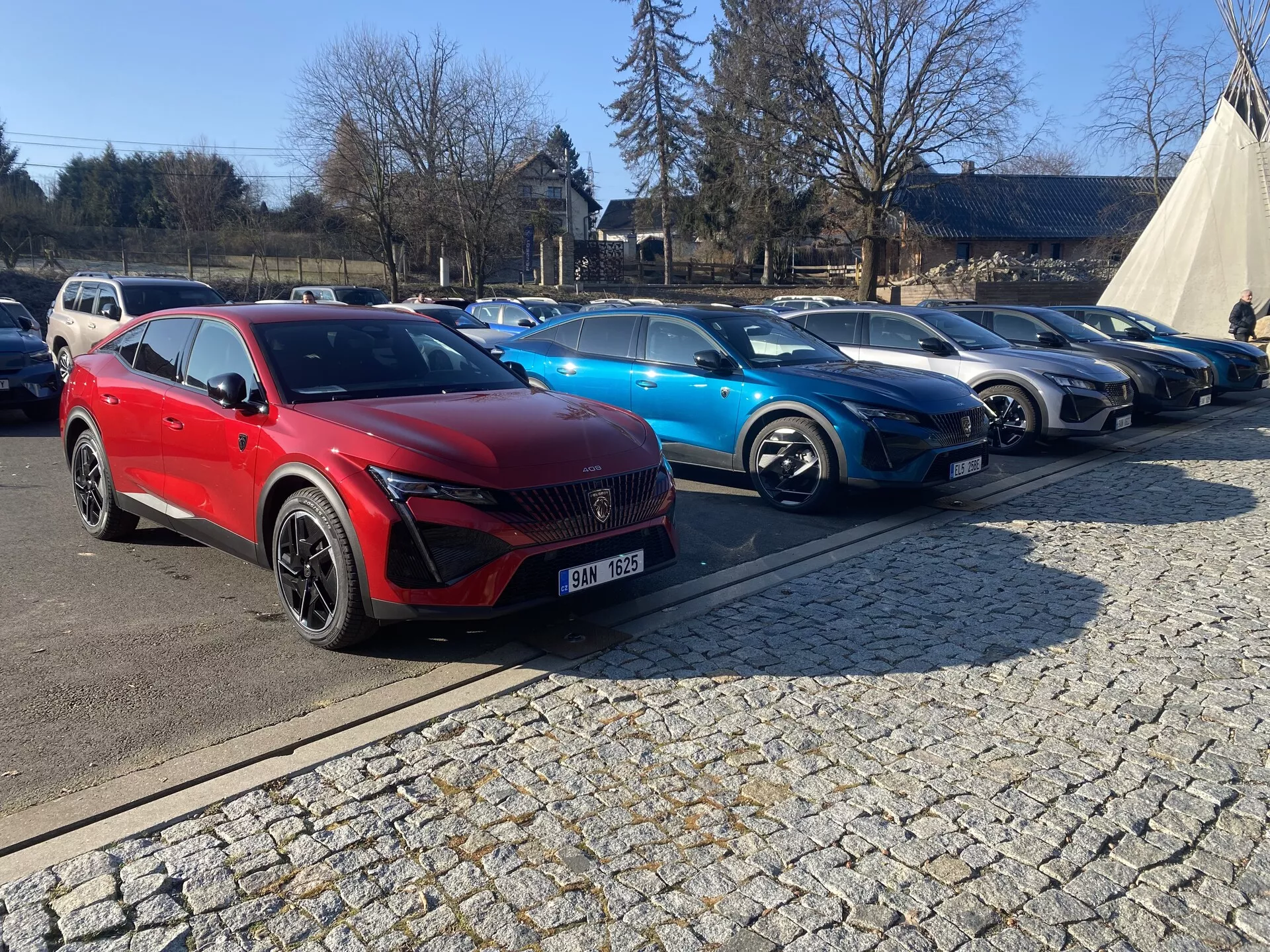 SUV rouge Peugeot 408 première génération 2023 présenté en plein jour lors d'un salon de l'automobile en plein air, mettant en avant son design angulaire moderne, sa grande calandre en motif de diamant, ses phares LED élégants et ses jantes en alliage sportives, avec d'autres voitures contemporaines et une tente promotionnelle en arrière-plan sous un ciel dégagé.
