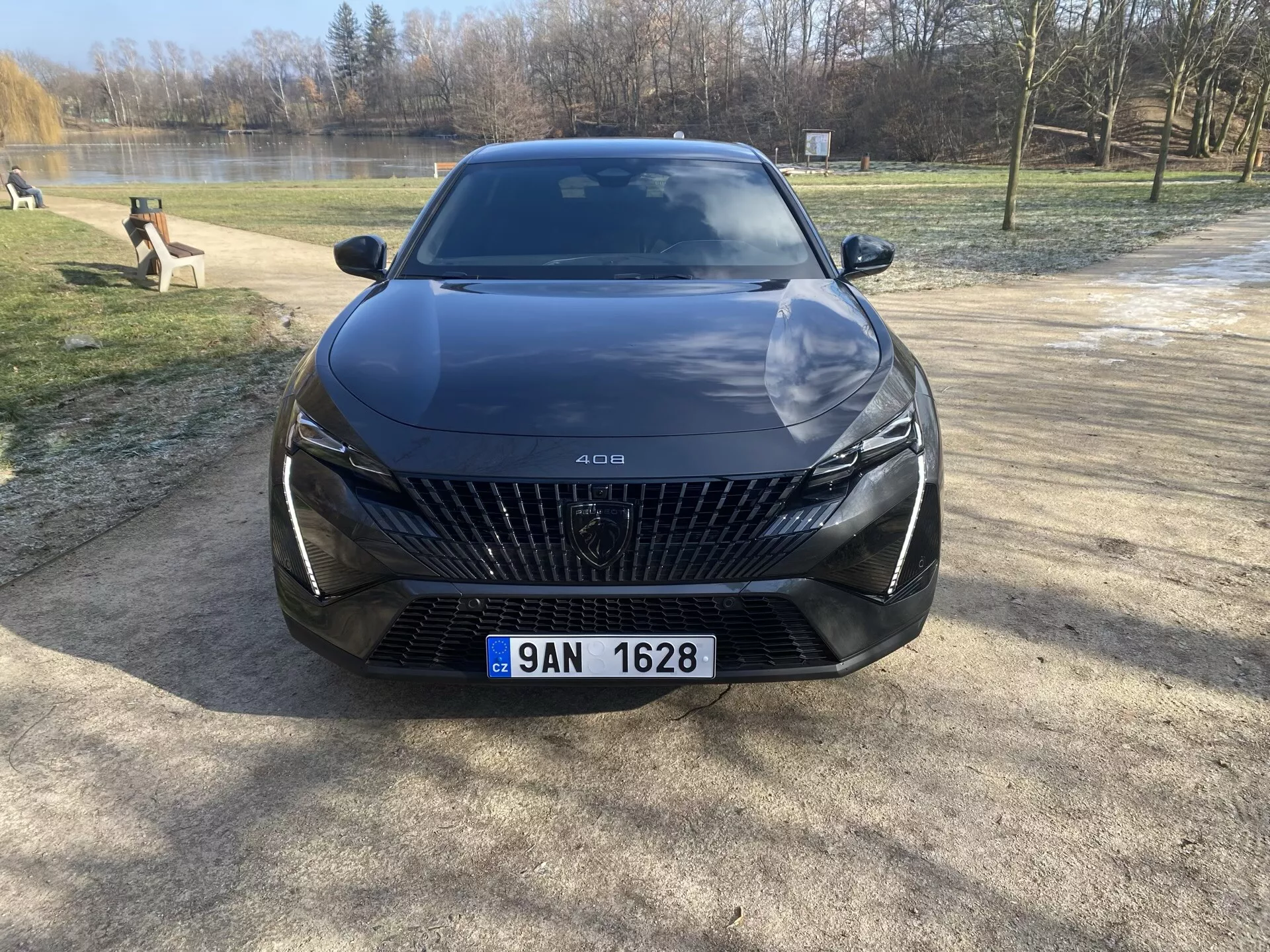 Vue de face de la Peugeot 408 2023 garée sur un sol gravillonné avec des arbres et un plan d'eau en arrière-plan, mettant en évidence sa grande grille avec des motifs en forme de diamant, phares LED élégants, lignes sculptées sur le capot et fonctionnalités avancées d'assistance au conducteur