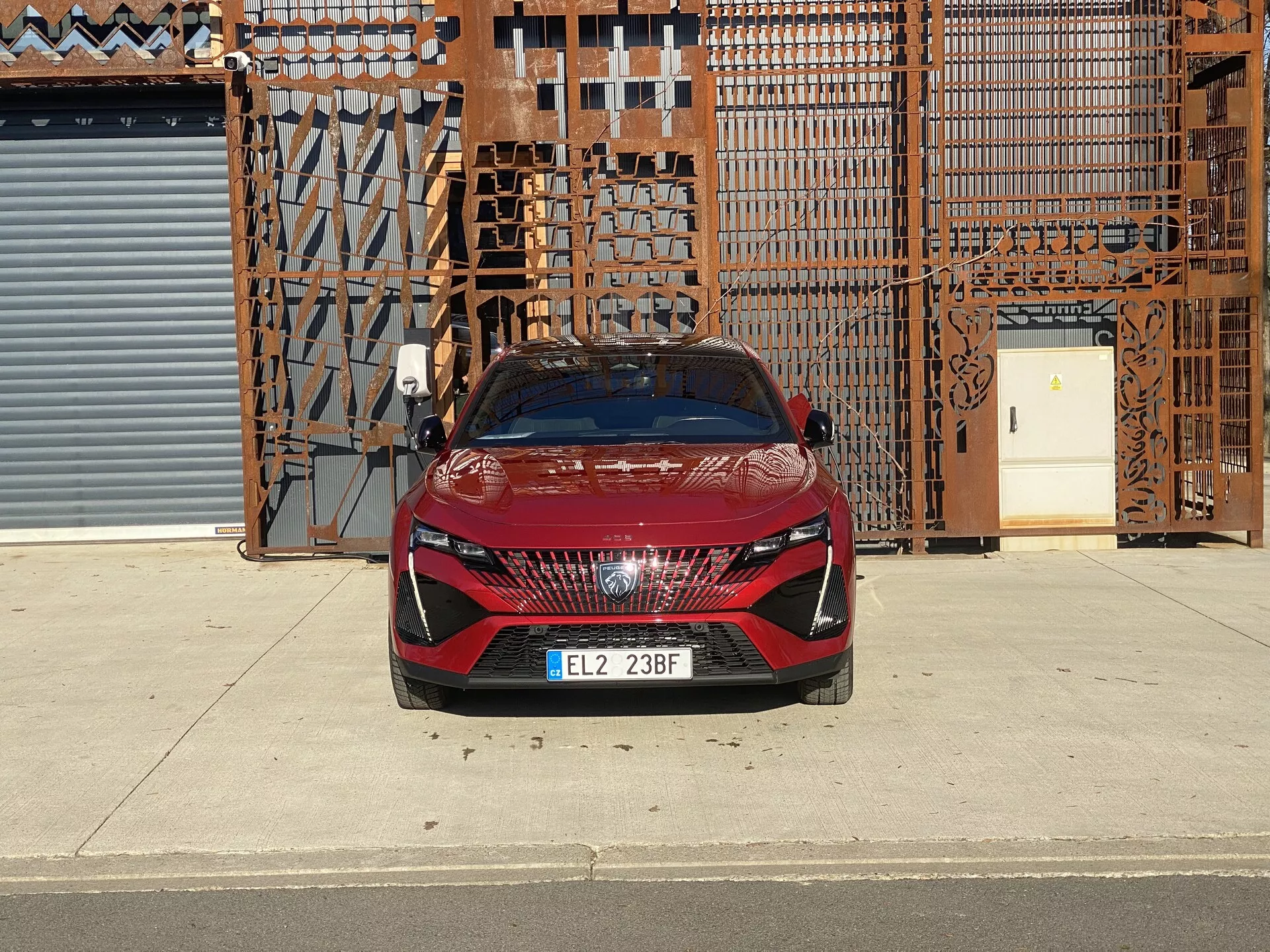 Vue frontale d’une Peugeot 408 de 2023 avec une peinture rouge et des accents noirs, mettant en avant une large grille avec un motif unique, des phares LED angulaires élégants et des lignes sportives sur le capot
