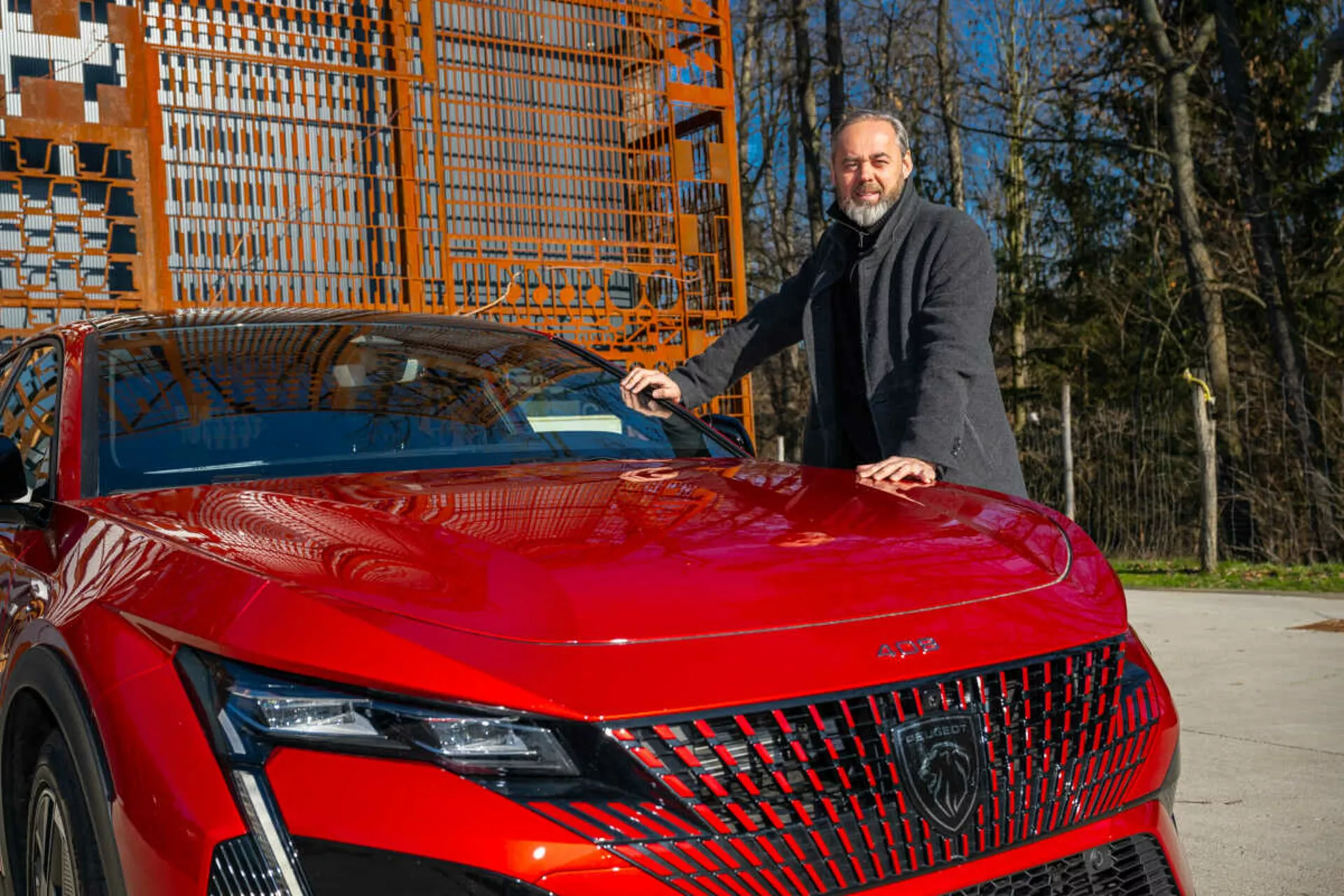 Modèle rouge de la Peugeot 408 2023 montrant son quart avant-gauche à la lumière du jour, mettant en évidence les phares LED détaillés, la grille géométrique et le capot sportif et aérodynamique avec un homme en manteau sombre à côté plaçant sa main sur le capot