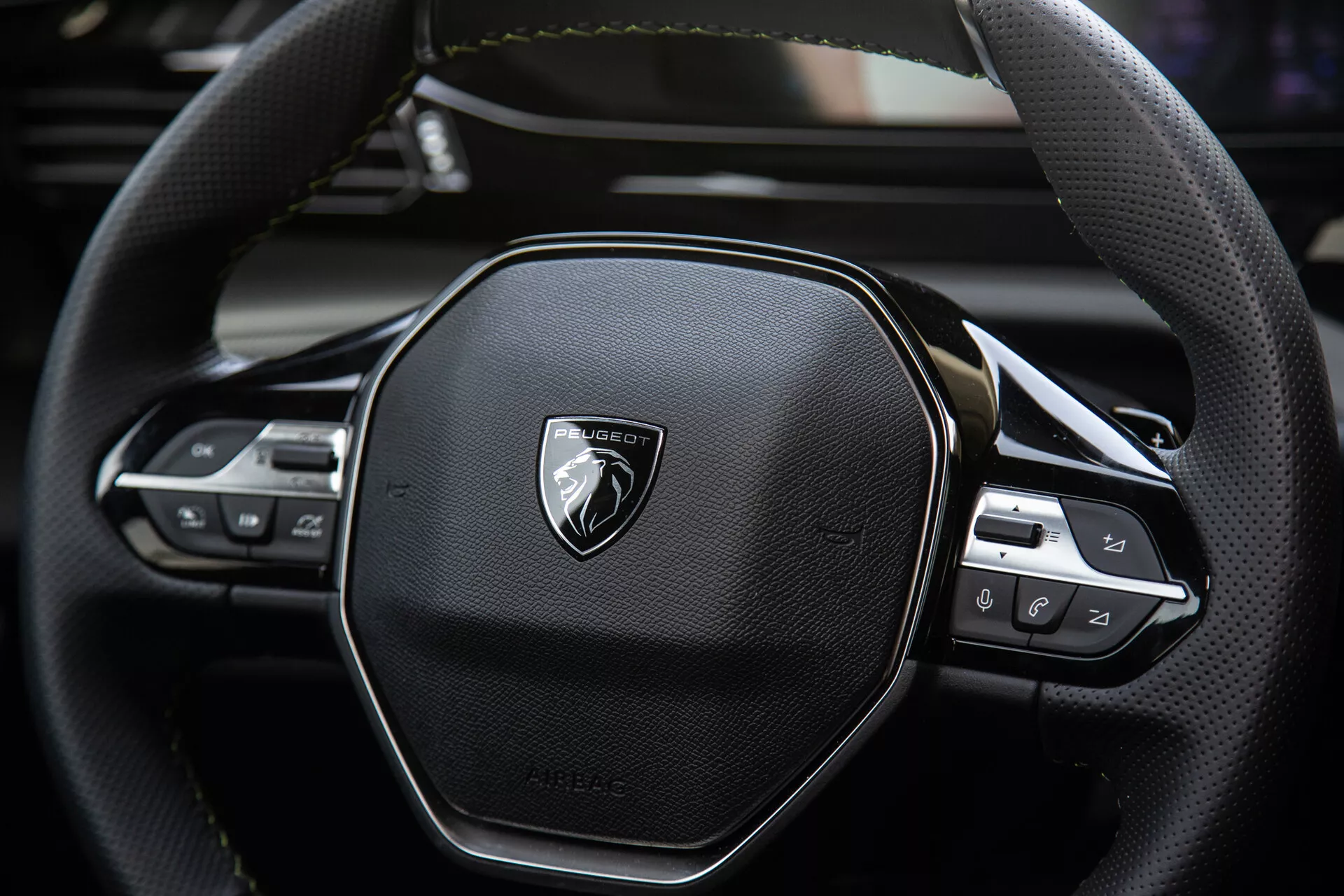 Vue rapprochée du volant du Peugeot 408 2023 présentant un design ergonomique hexagonal, couture en cuir, commandes technologiques intégrés pour audio et réglages de croisière, avec emblème sur métal brossé.