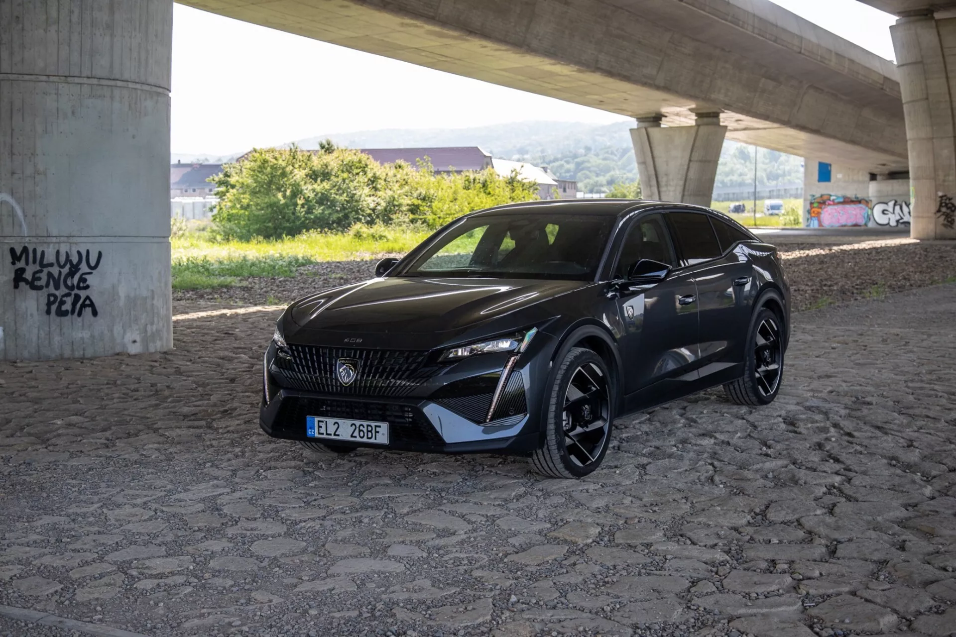 Peugeot 408 de 2023 capturée sous un angle de trois quarts mettant en évidence ses phares LED élégants, sa grille à motif et son pare-chocs avant sportif aérodynamique sous la lumière naturelle du jour dans une zone pavée sous un viaduc