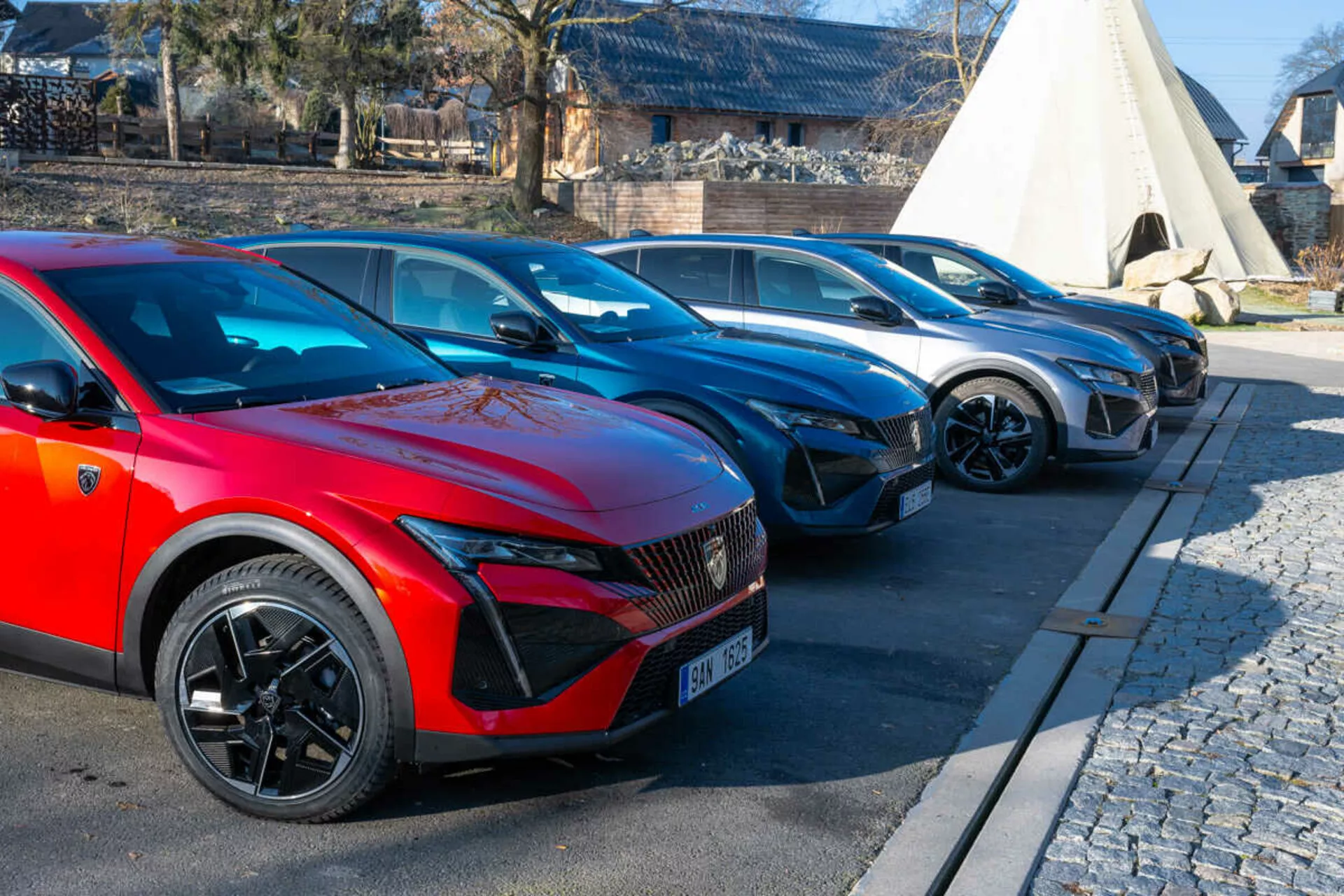 Gamme Peugeot 408 2023 présentant une voiture rouge au premier plan avec des roues sport à multiples branches et des phares LED indicatifs de modèles électriques ou hybrides, garée sur une zone pavée à côté de pavés