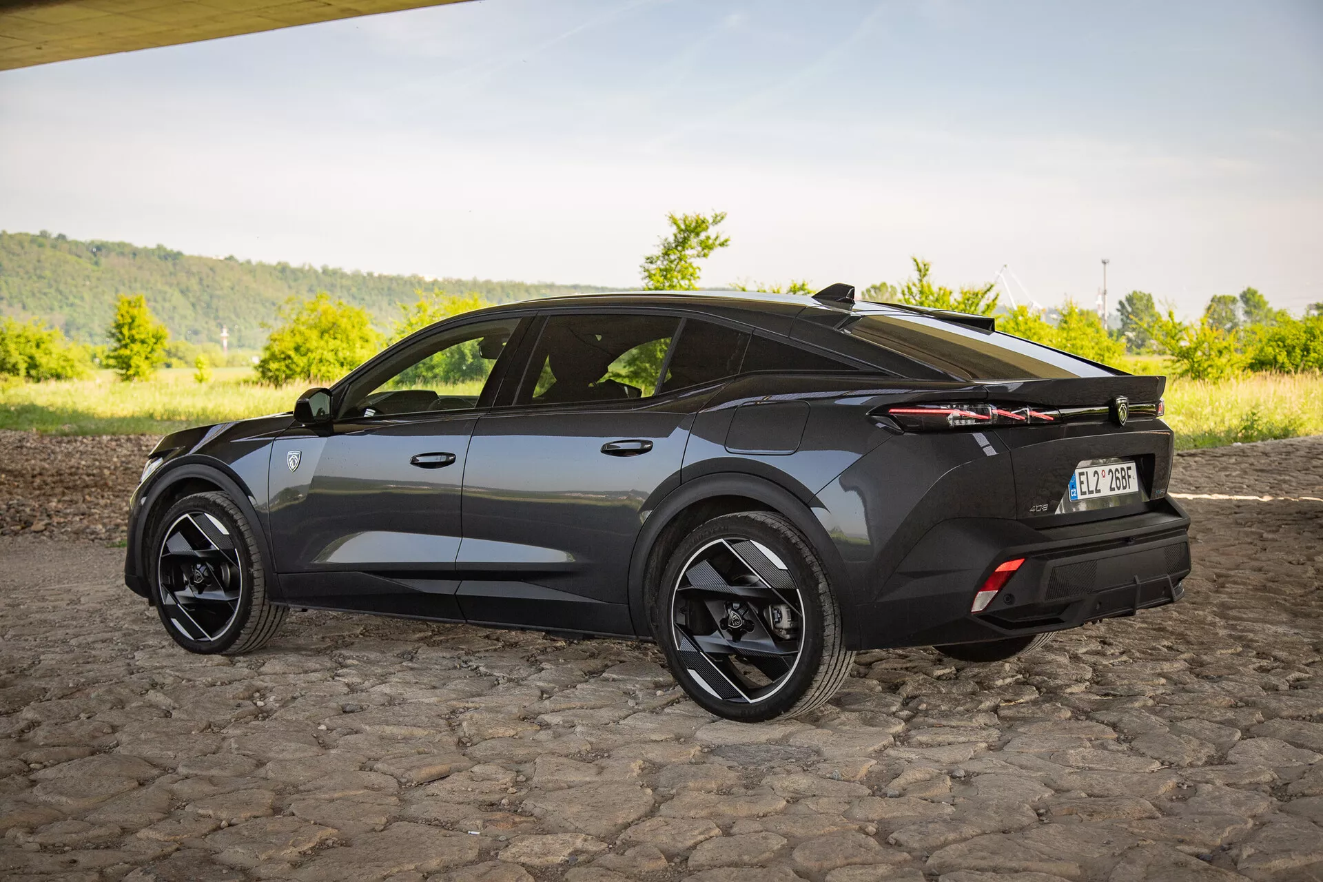 SUV crossover Peugeot 408 de 2023 en finition sombre, capturant une vue arrière aux trois quarts mettant en lumière un design aérodynamique avec des angles marqués, une ligne de toit sportive inclinée, de grandes roues en alliage noir, des feux arrière LED distinctifs et un spoiler arrière subtil sur une surface en pavés