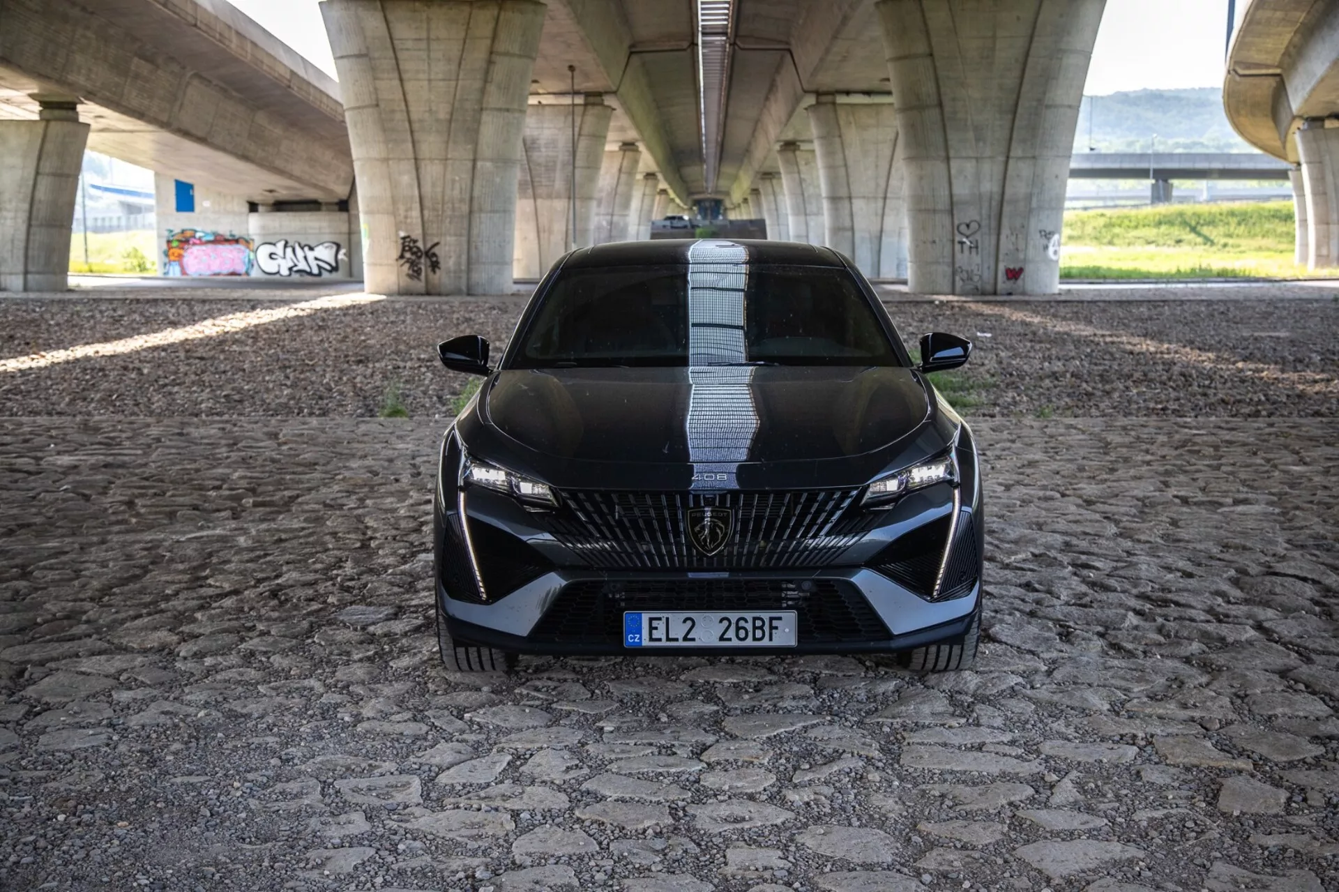 Peugeot 408 2023 dans une vue frontale dynamique mettant en avant sa position agressive et un design sportif avec une grille détaillée, des phares LED élégants et des caractéristiques aérodynamiques sur un pavage à motif sous un pont urbain