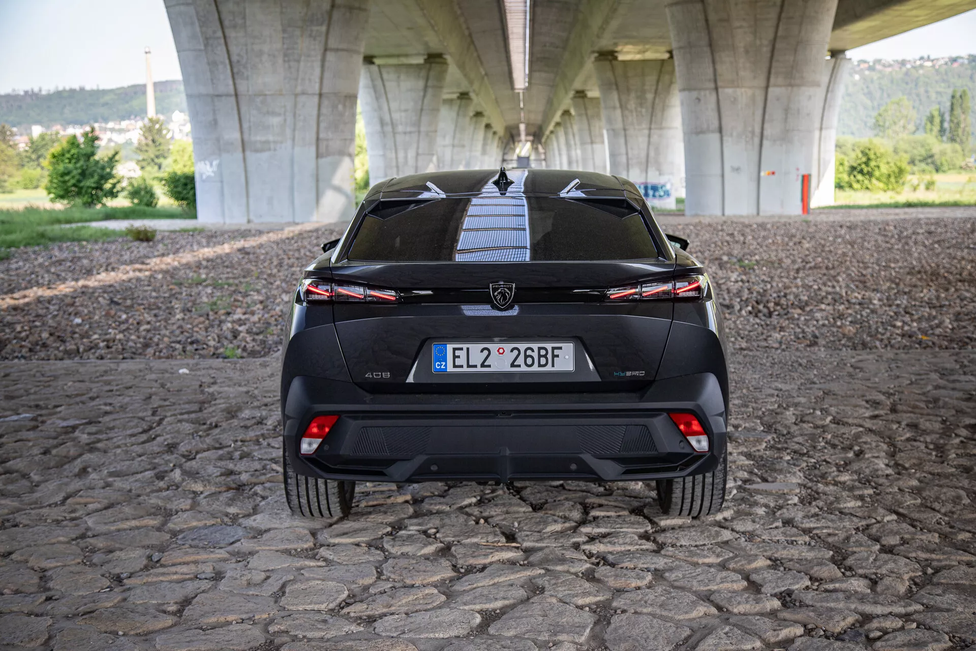 Vue arrière de la Peugeot 408 2023 présentant le hayon, le pare-chocs, le diffuseur inférieur, les feux arrière reliés par une barre lumineuse et les capteurs d'aide au stationnement, photographiée sous un pont sur une surface pavée
