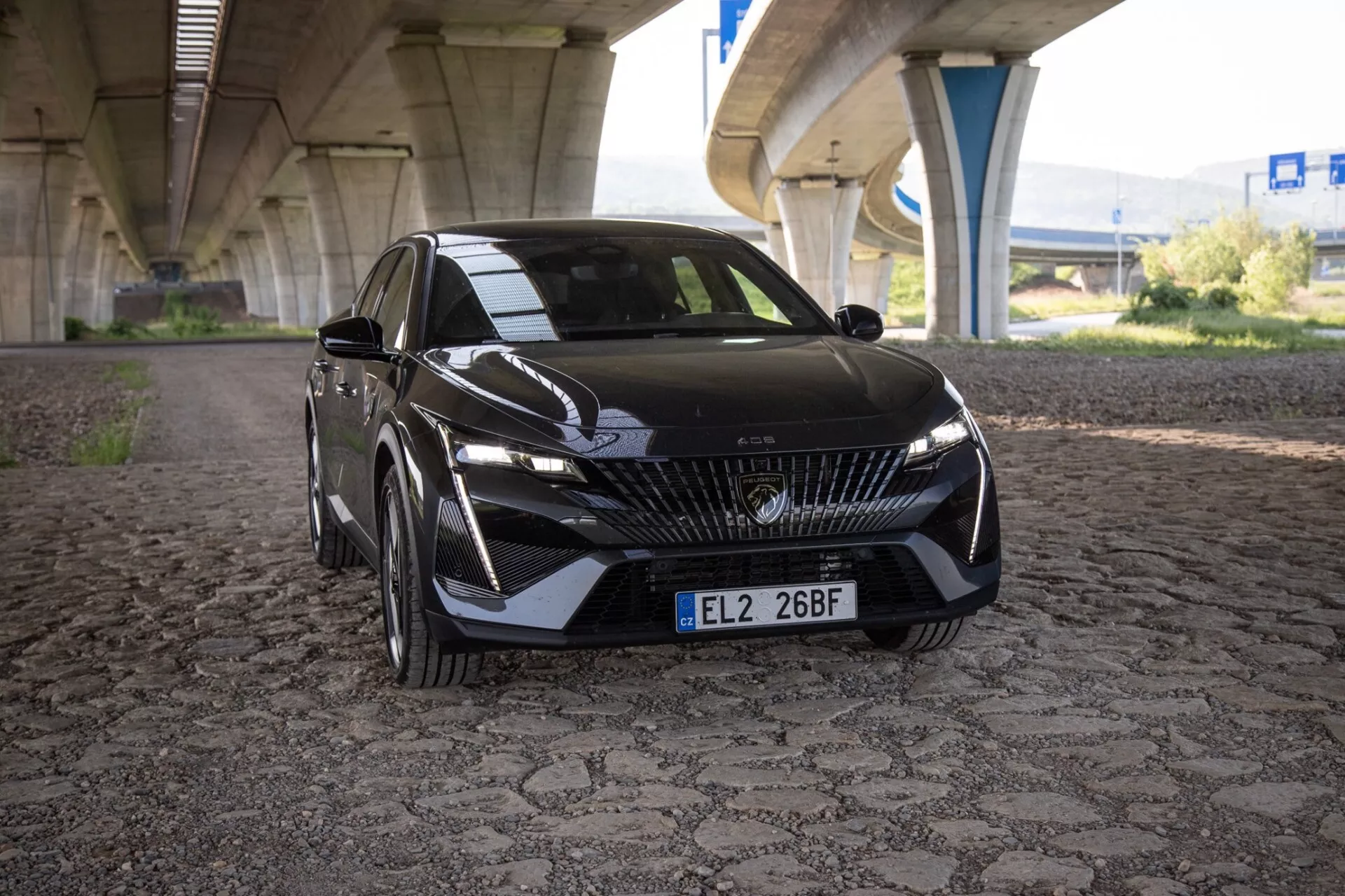 Vue frontale légèrement décentrée de la Peugeot 408 2023 montrant le design avant détaillé incluant la grille, le logo, les phares LED, le pare-chocs, les prises d'air, les contours du capot et la plaque d'immatriculation avant, sur un fond urbain avec pont.