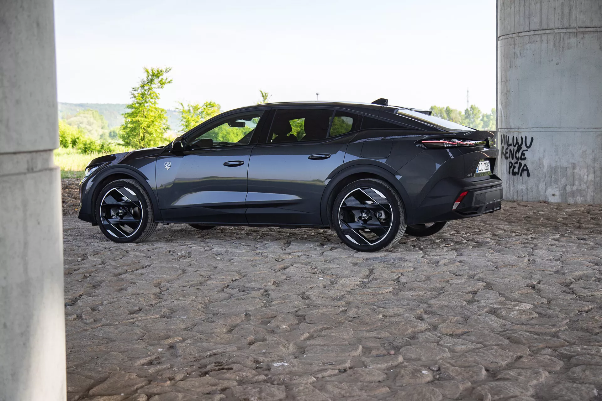 Vue de profil d'une voiture moderne de couleur sombre Peugeot 408 2023 sous un pont avec graffiti, mettant en avant un design aérodynamique, des jantes en alliage et des vitres teintées