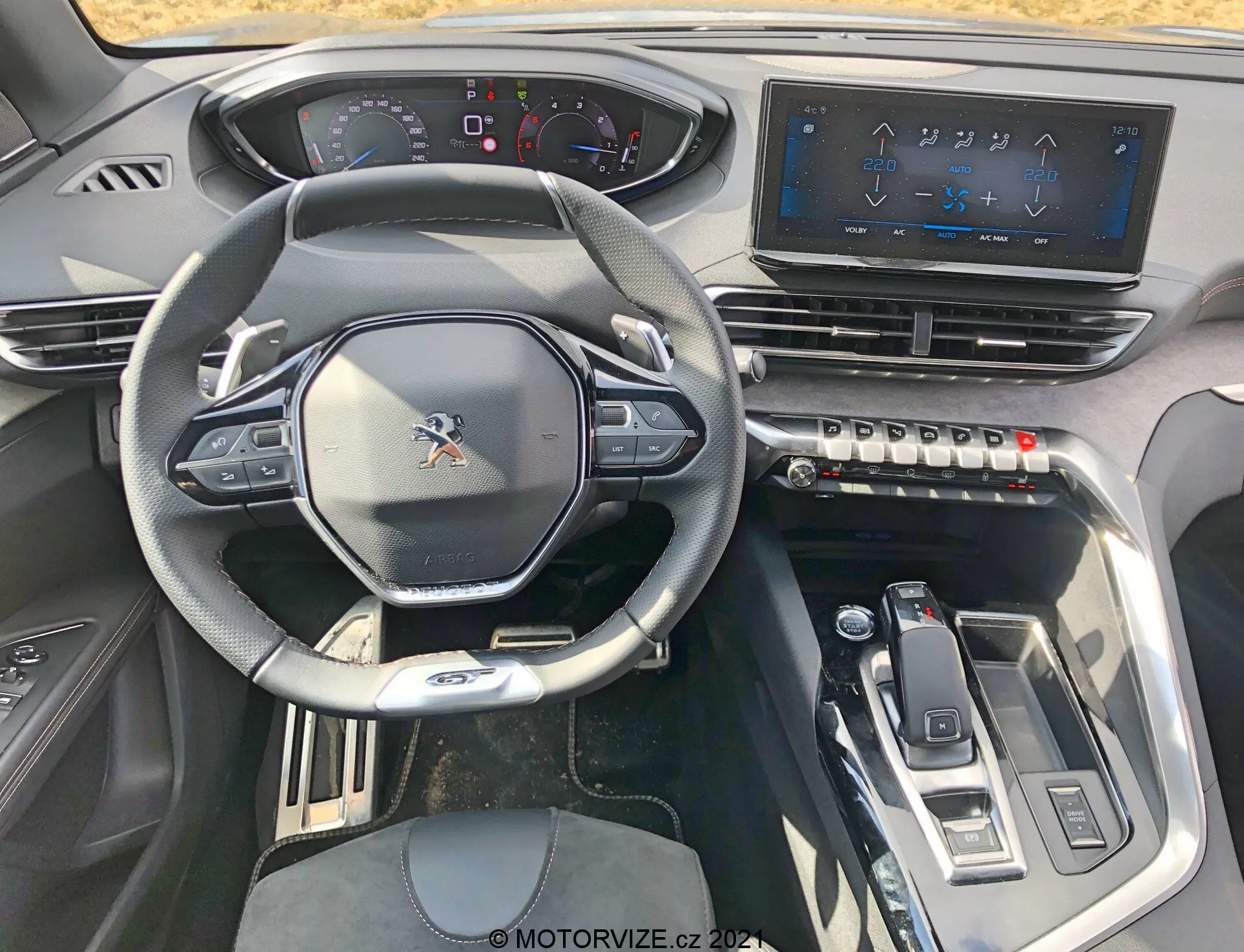 Vue intérieure d'une Peugeot 5008 2021 depuis le siège du conducteur, montrant le volant multifonction sportif avec commandes intégrées, un tableau de bord partiellement numérique, un grand système d'infodivertissement à écran tactile, des boutons de contrôle tactile pour la climatisation, un sélecteur de vitesse élégant et des éléments de design modernes avec des accents métalliques pour une expérience high-tech et haut de gamme.