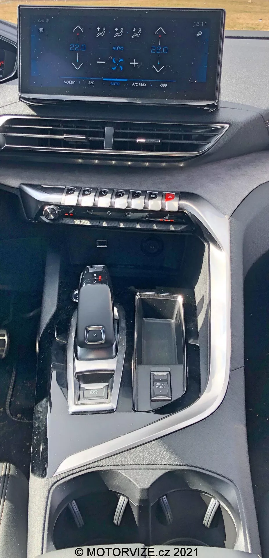 Vue intérieure de la Peugeot 5008 de 2021 montrant la console centrale vue d'en haut. L'image met en avant un écran tactile flottant d'infodivertissement affichant les commandes de la climatisation au-dessus de boutons et de cadrans physiques pour le contrôle du climat. Plus bas, un levier de vitesses électronique élégant et les commandes environnantes pour la sélection des modes de conduite et un bouton d'interface multifonctionnelle sont visibles. Le design indique un véhicule moderne et haut de gamme avec des fonctionnalités technologiques avancées et ergonomiques, incluant des porte-gobelets et des espaces de rangement.