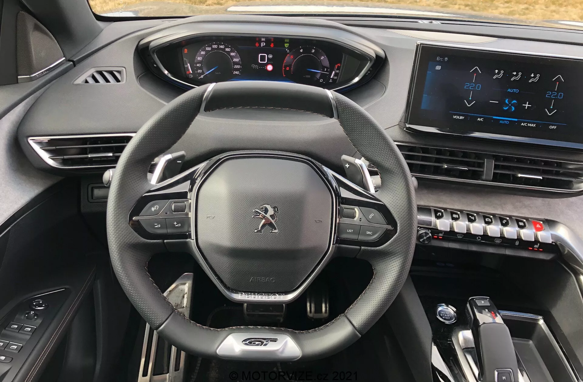Vue intérieure de la Peugeot 5008 2021 depuis le siège du conducteur, mettant en avant le volant gainé de cuir à fond plat avec commandes intégrées, un tableau de bord entièrement numérique avec indicateurs lumineux, un grand écran tactile d'infodivertissement sur la console centrale, des commandes de climatisation élégantes, un sélecteur de vitesses électronique pour transmission automatique et un design de matériaux sombres de haute qualité évoquant une esthétique moderne, high-tech et premium.