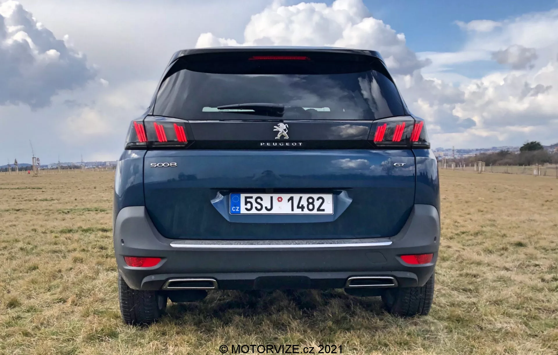 Vue arrière d'un SUV Peugeot 5008 moderne, mettant en évidence les feux arrière horizontaux élégants avec des segments LED en forme de griffes distinctifs, le logo Peugeot et l'inscription du modèle '5008'. Couleur bleu foncé ou gris, garé sur un terrain extérieur avec un ciel partiellement nuageux. Les éléments visibles comprennent le pare-brise arrière, l'essuie-glace arrière, le feu stop monté en hauteur, la poignée de hayon et la plaque d'immatriculation de la République tchèque avec 'CZ'. Bande réfléchissante au-dessus du pare-chocs avec des capteurs de stationnement et des découpes d'embouts d'échappement évidentes. Prise de vue centrée avec la voiture remplissant la majorité du cadre.
