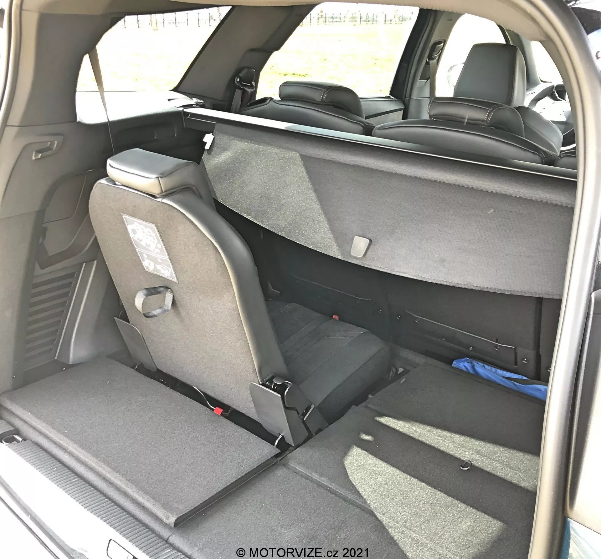 Vue intérieure de l'aire de chargement d'une Peugeot 5008 2021 sous un angle arrière trois quarts droit, montrant une disposition des sièges flexible avec les sièges arrière partiellement rabattus, sellerie en tissu, appuie-têtes et ceintures de sécurité. Siège arrière droit complètement rabattu, siège gauche en position verticale, indiquant une fonctionnalité de rabattage fractionné 60/40. Garniture intérieure sombre visible avec une illumination naturelle à la lumière du jour, prise par MOTORVIZE.cz.