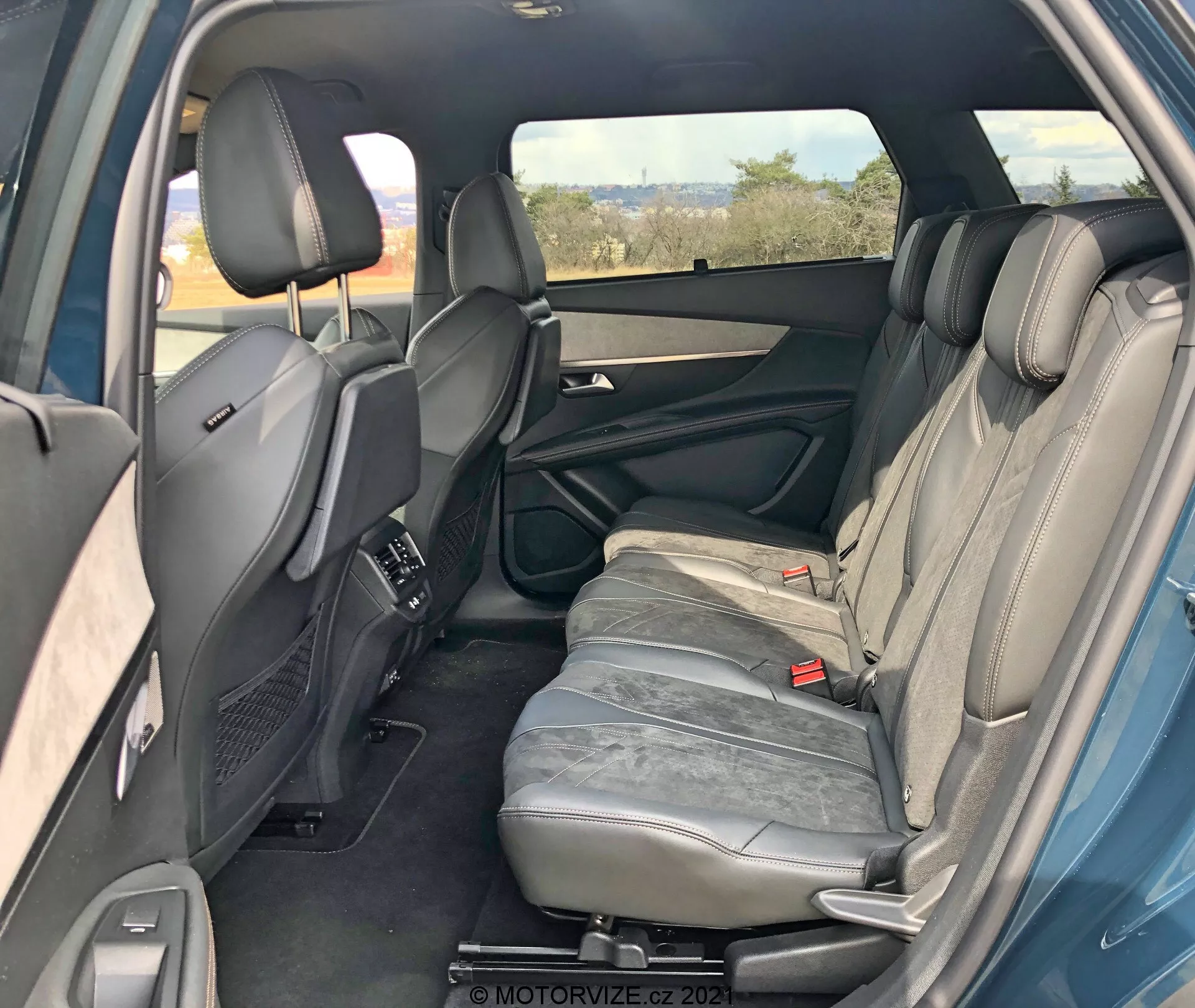 Intérieur d'une Peugeot 5008 2021 vu de l'arrière droit montrant le compartiment passager arrière. La porte arrière droite est ouverte révélant les caractéristiques du panneau de porte comme la poignée, les compartiments de rangement et le haut-parleur. Trois sièges arrière en cuir gris avec appuis-tête sont visibles. L'accoudoir du siège central est en position verticale. Les ceintures de sécurité arrière et les sièges avant avec appuis-tête réglables et console centrale sont également visibles. Bouches de contrôle du climat arrière sur la console centrale, fenêtre droite teintée, aperçus de la porte arrière gauche indiquant la symétrie. Un intérieur bien entretenu sans caractéristiques technologiques visibles.