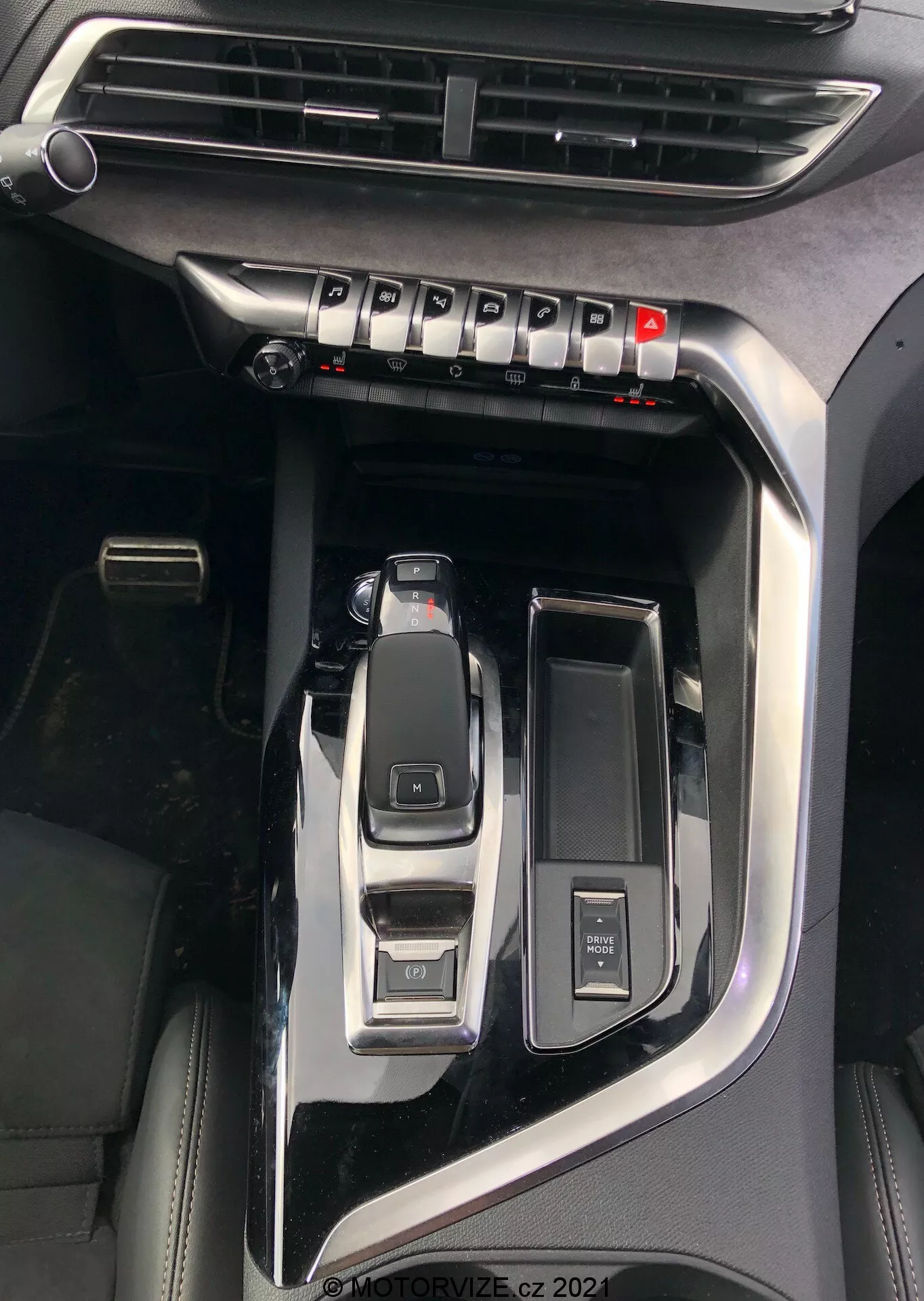 Vue rapprochée de la console centrale de la Peugeot 5008 2021 mettant en évidence des caractéristiques clés telles que le levier de vitesses automatique en position de stationnement, le sélecteur de mode de conduite pour les modes éco, sport, confort, un bouton de démarrage/arrêt pour l'allumage sans clé, une molette de contrôle multimédia, un porte-gobelet ouvert, une bouche d'aération visible pour le contrôle du climat et une finition en noir piano avec des accents métalliques indiquant un modèle premium, filigrane 'MOTORVIZE.cz 2022'.