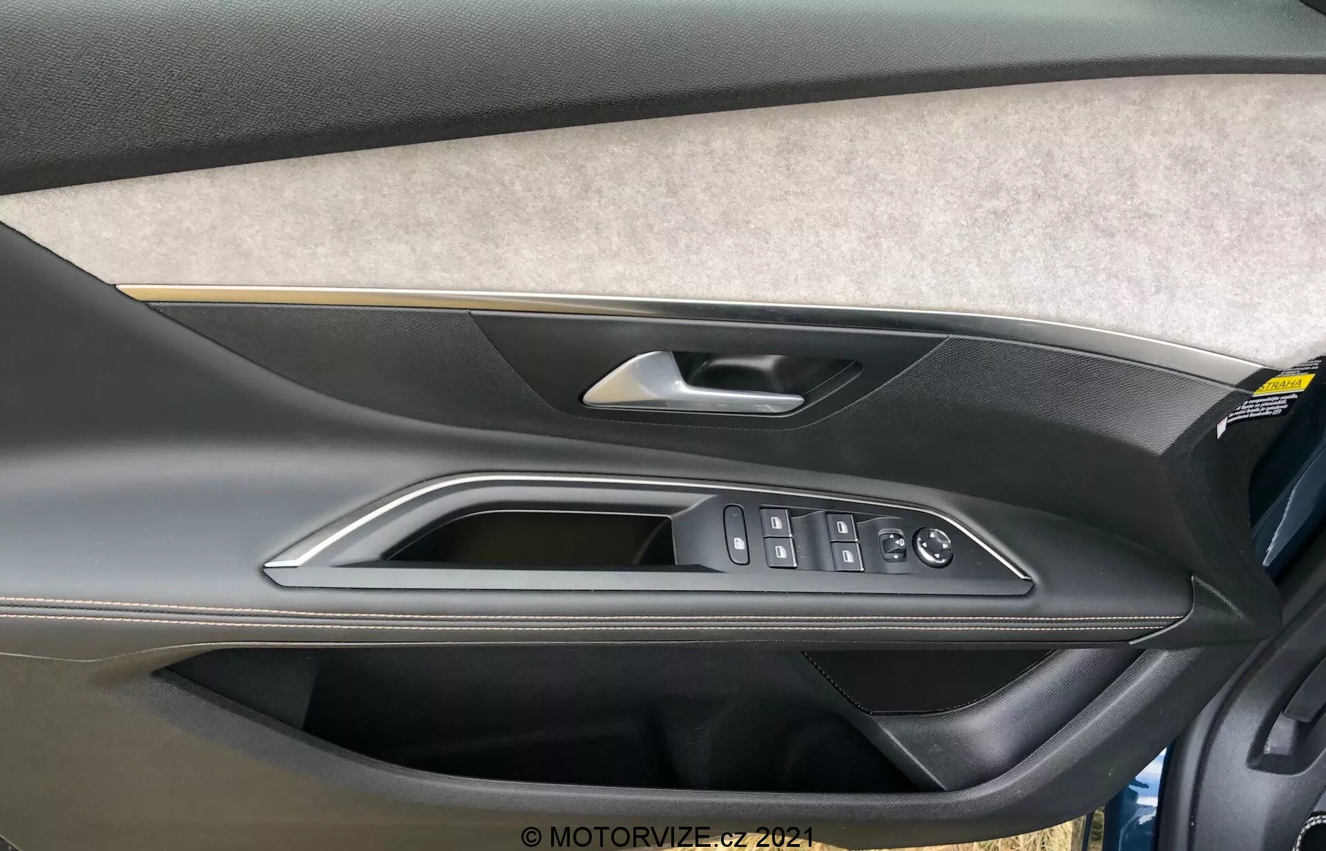 Vue rapprochée du panneau de porte intérieur de la Peugeot 5008 2021 avec une section revêtue de feutre ou de tissu, un design en plastique moulé bicolore avec une partie supérieure plus sombre et une partie inférieure plus claire, une poignée de porte en matériau métallique ou chromé de haute qualité, des garnitures polies et des commandes ergonomiques pour les fenêtres électriques, la serrure de porte et les rétroviseurs latéraux. Une poche de rangement est également visible en bas du panneau de porte.