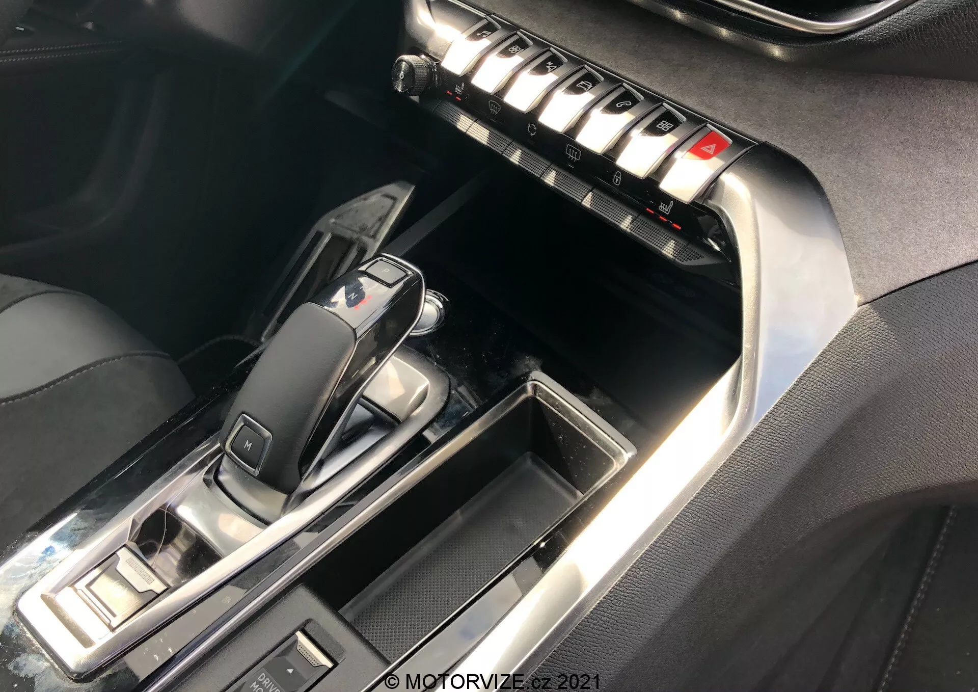 Gros plan sur la console centrale et l'intérieur avant de la Peugeot 5008 2021, se concentrant sur la zone du levier de vitesses automatique et la pile centrale. Le levier de vitesses, en position 'P', présente un design élégant avec du plastique noir brillant et des garnitures métalliques. Les boutons environnants suggèrent des modes de conduite et des commandes de frein de stationnement. La pile centrale comprend une rangée de boutons métalliques probablement pour le contrôle climatique, les dégivreurs et les réglages des sièges. Les surpiqûres contrastées sur le cuir ou le similicuir et les accents chromés rehaussent la sensation de luxe. L'éclairage met en valeur les surfaces réfléchissantes et les détails.