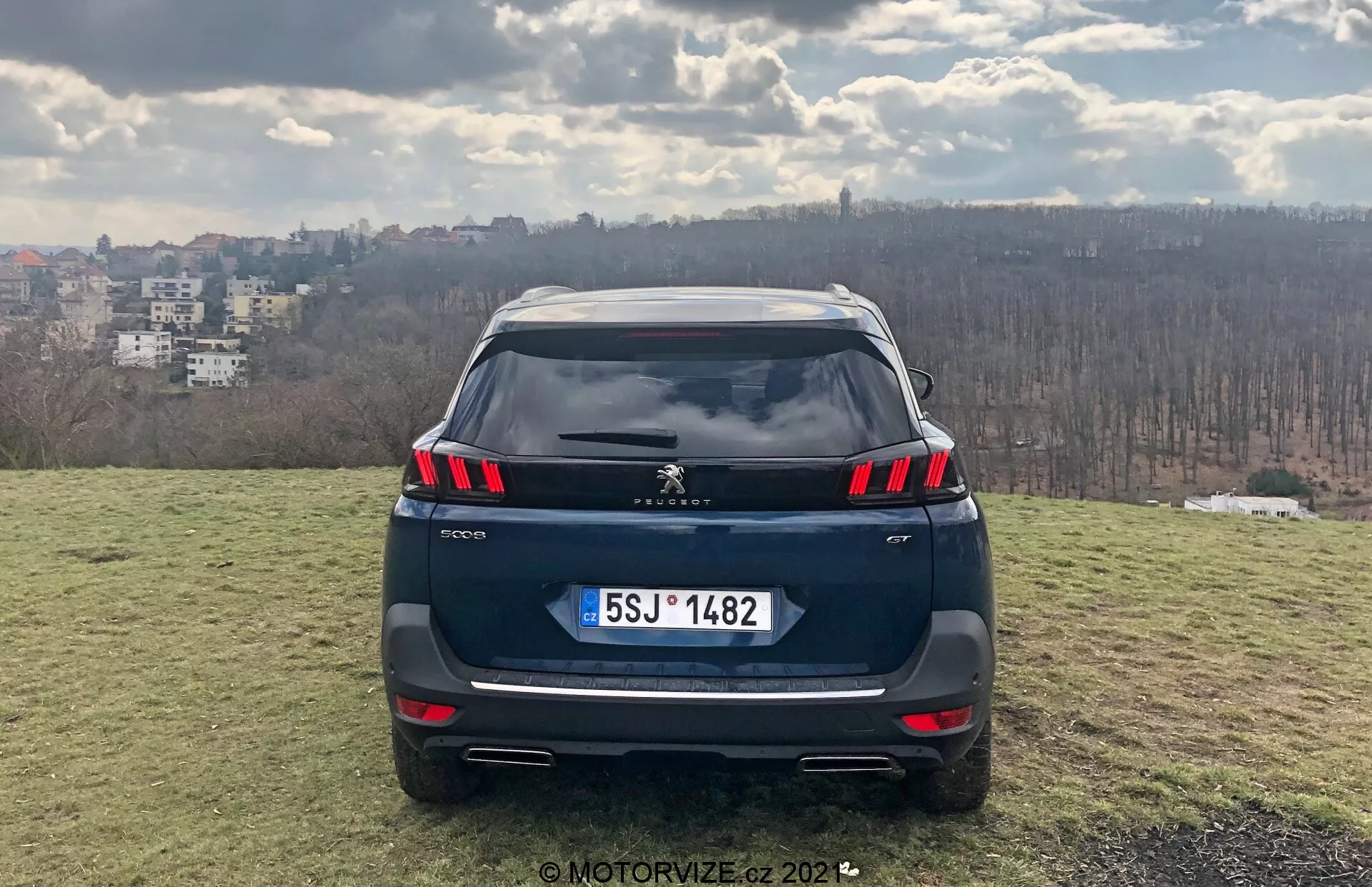 Vue arrière face d'un SUV Peugeot 5008 2021 sur une zone herbeuse surélevée avec vue sur la ville en arrière-plan, présentant des feux arrière LED en griffe, capteurs de stationnement intégrés dans le pare-chocs, possible caméra de recul, plaque d'immatriculation de style européen, doubles sorties d'échappement, spoiler de toit et antenne aileron de requin; un cadre de jour suggérant une partielle nébulosité, photographié par MotorVize.cz en 2022.