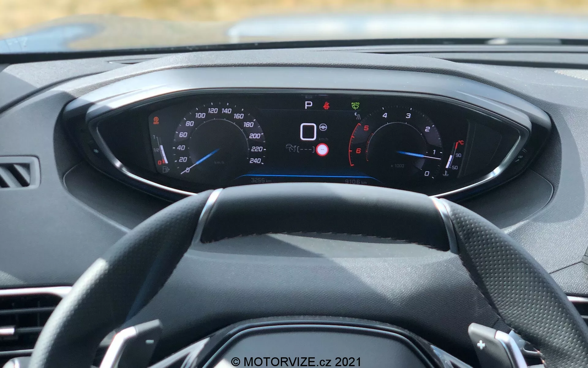 Vue intérieure d'un véhicule moderne depuis une perspective à la première personne, mettant l'accent sur le volant et le tableau de bord numérique. Le volant à trois branches est centré, avec divers cadrans et indicateurs sur le tableau de bord illuminé derrière lui. Le compteur de vitesse et le compte-tours sont visibles, avec des lectures en km/h et mph, adaptés aux marchés internationaux. Le tableau de bord affiche de nombreuses lumières d'avertissement et d'information, telles que frein de stationnement activé, portes ouvertes, rappel de ceinture de sécurité et faible pression des pneus, indiquant des fonctionnalités avancées de diagnostic et de sécurité à bord. Le schéma de couleurs du tableau de bord est bleu et rouge sur un fond noir avec du texte blanc. Cette image, prise à la lumière du jour, met en évidence le design intérieur de la voiture, l'instrumentation numérique et la technologie moderne.