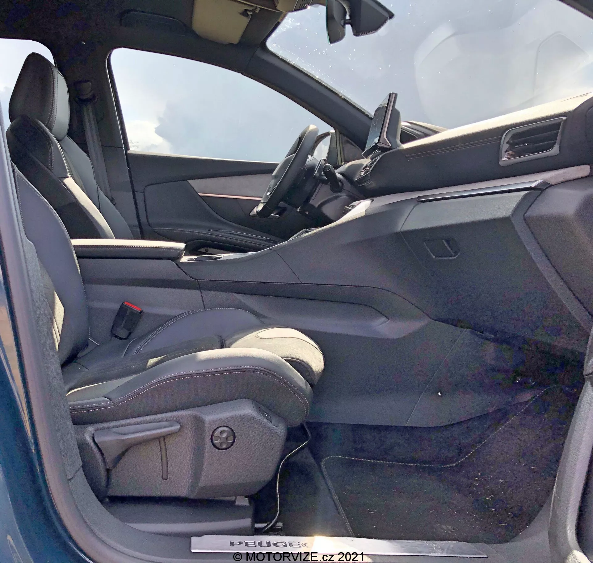Vue de l'intérieur d'une Peugeot 5008 de 2021 depuis le côté conducteur avec la porte ouverte. Le siège conducteur est de style baquet, probablement en cuir ou en matériau similaire, avec des renforts latéraux et des commandes de réglage électrique. La porte du côté conducteur ouverte montre un panneau intérieur avec des commandes de vitres électriques, probablement des commandes de rétroviseurs et des mécanismes de verrouillage de la porte. Le tableau de bord comprend un écran tactile d'infodivertissement central avec un design minimaliste. La partie supérieure du volant est visible, suggérant un volant en cuir avec des commandes intégrées. L'intérieur présente un schéma de couleurs bicolore avec des surfaces supérieures plus sombres et des surfaces inférieures plus claires.