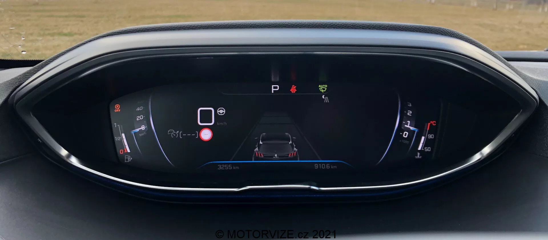 Tableau de bord du Peugeot 5008 2021, affichant un écran numérique vu depuis le siège du conducteur. L'écran comprend un indicateur de puissance/batterie, un graphique central de la voiture avec des icônes d'avertissement, un indicateur 'P' de stationnement, un indicateur de carburant et des informations sur l'odomètre/le voyage. Le design présente un schéma de couleurs moderne bleu et rouge sur un boîtier incurvé et élégant. Filigrane © MOTOR1ZED 2021.