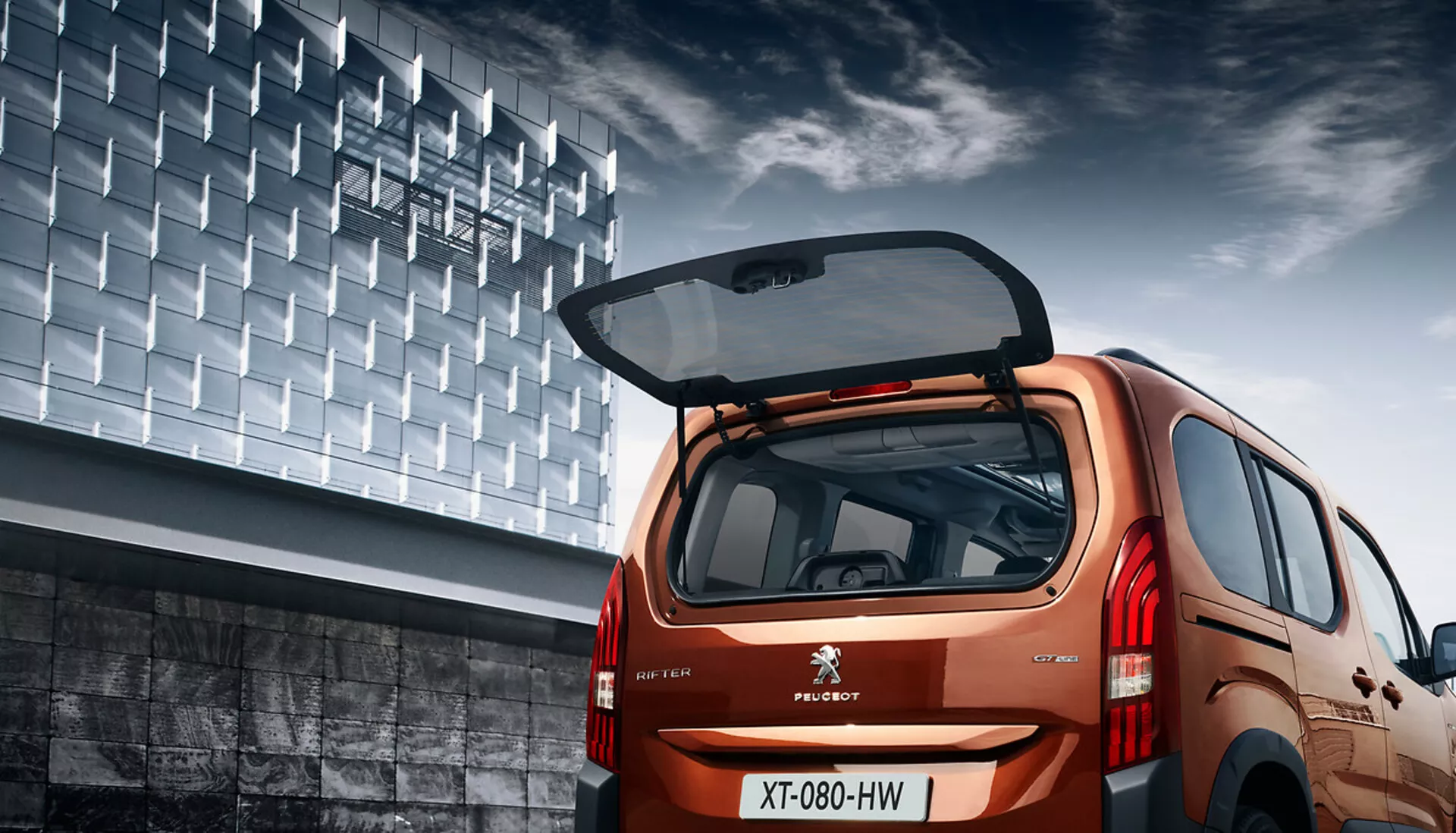 Vue arrière en angle du Peugeot Rifter 2018 montrant son design hatchback, finition métallique, hayon ouvert avec espace de chargement visible, feux arrière LED et décor urbain.