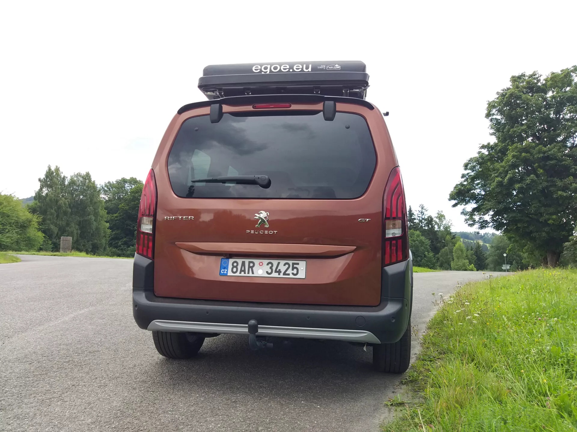 Vue arrière d'un Peugeot Rifter 2018, montrant le pare-brise arrière, les feux arrières, le pare-chocs et la plaque d'immatriculation, avec le logo Peugeot et le nom du modèle Partner visibles. La voiture, garée devant un fond naturel, est équipée d'une boîte de toit et arbore un accessoire de marque 'egoe.eu', en couleur rouge/maroon.