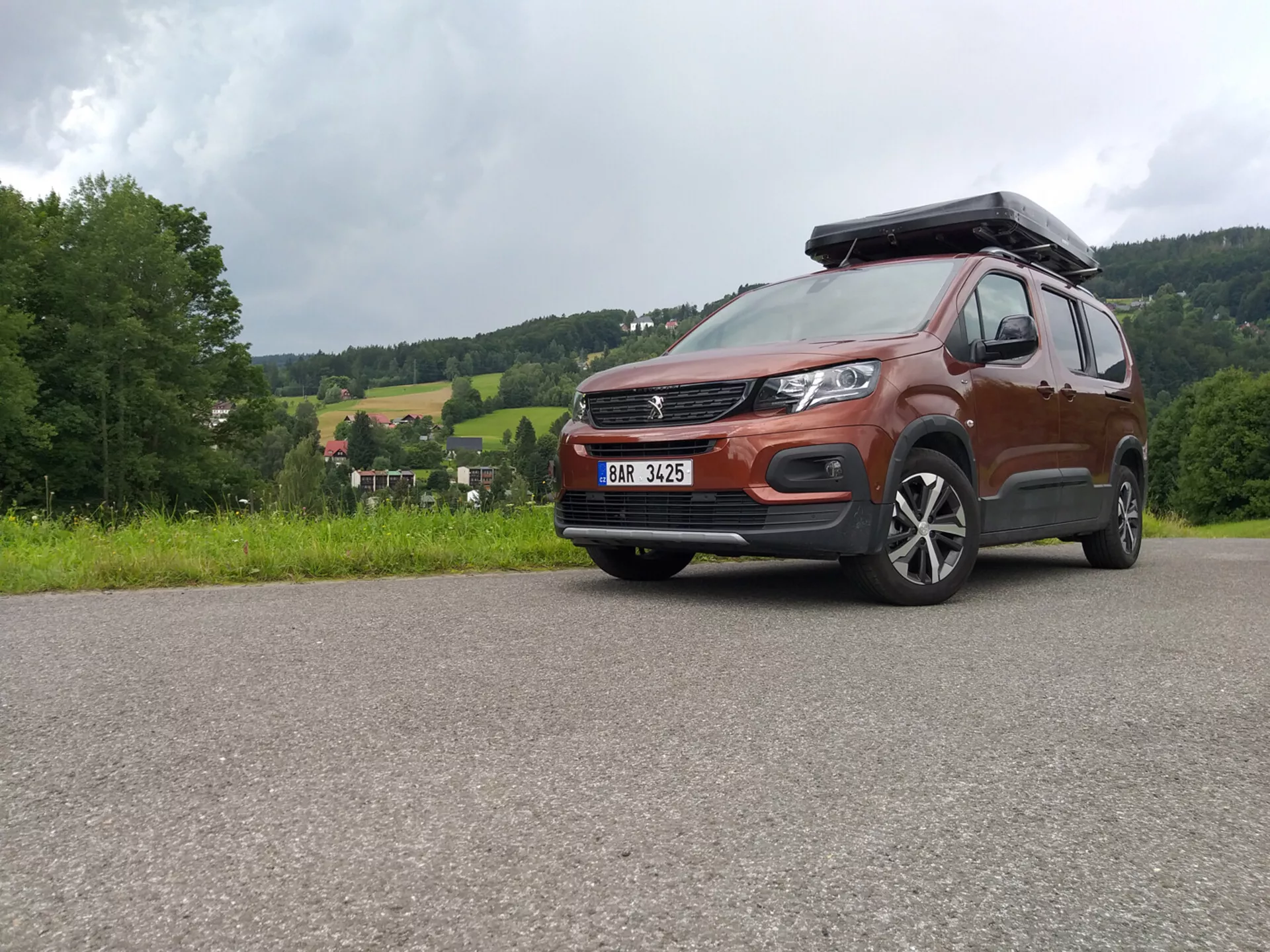 SUV Peugeot Rifter 2018 avec peinture métallique et coffre de toit capturé sous un angle bas mettant en évidence la grille, le pare-chocs et les roues en alliage, montrant les capacités tout-terrain et le design moderne incluant des phares LED et des systèmes d'assistance au conducteur ADAS, stationné sur une surface pavée contre un paysage rural