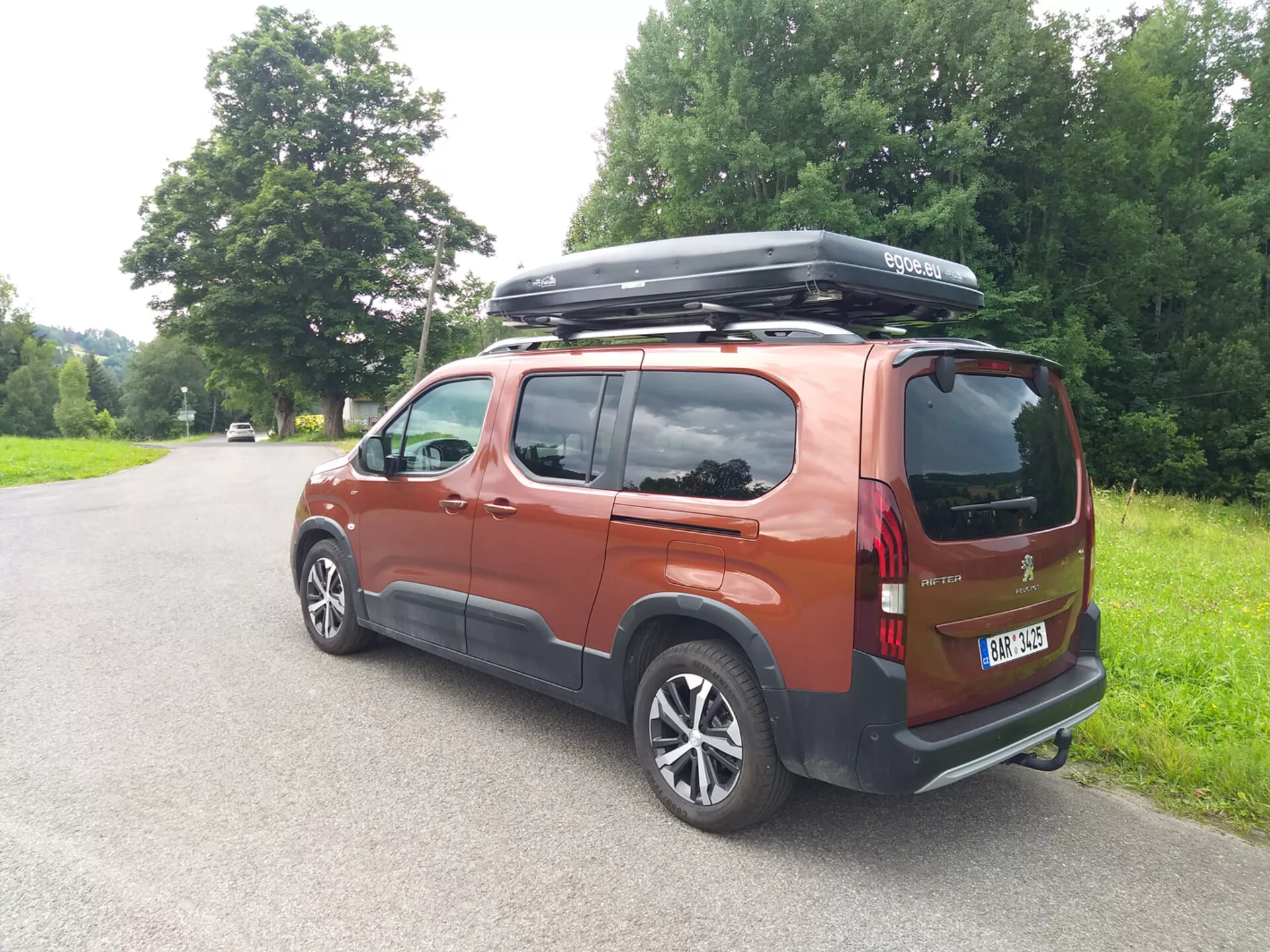 Vue arrière trois quarts d'un Peugeot Rifter 2018 en orange brûlé métallisé garé sur une route rurale, équipé de jantes en alliage, feux arrière LED, box de toit, vitres teintées et une courte antenne sous un éclairage naturel de jour