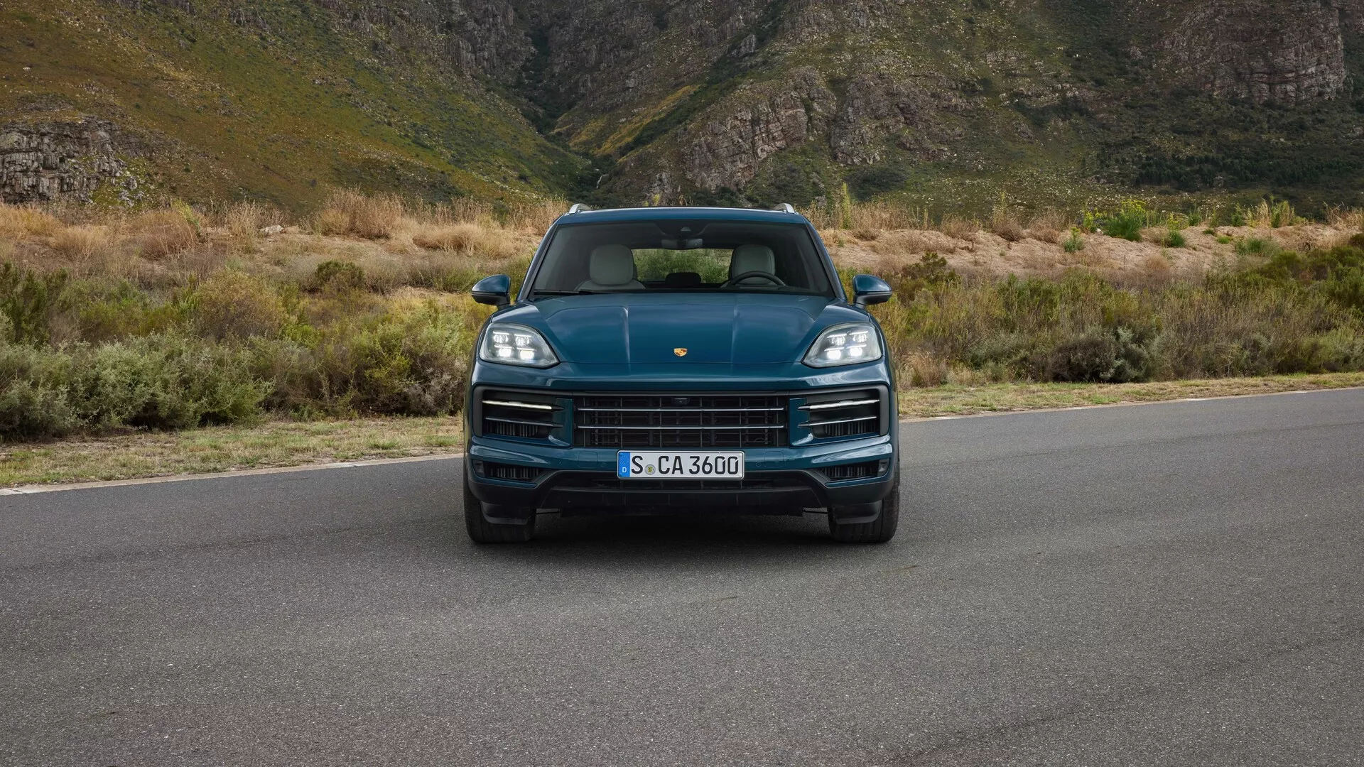 Porsche Cayenne 2023 (9YA Facelift) vue de face au niveau des yeux mettant en avant les phares LED, la calandre proéminente avec le logo de la marque, le design sportif du capot et les capteurs de stationnement intégrés, sur un fond montagneux indiquant luxe et aventure