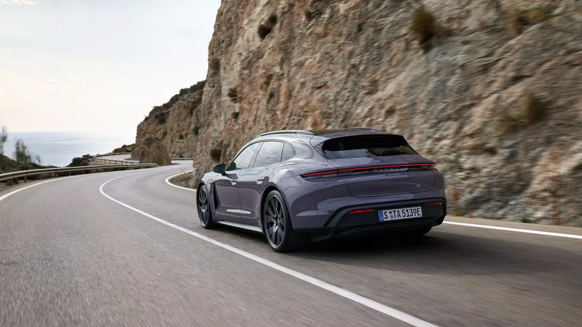 Vue arrière dynamique trois quarts de la Porsche Taycan 2024 Y1A facelift sur une route côtière, mettant en évidence la posture sportive, la silhouette profilée et les caractéristiques technologiques avancées.