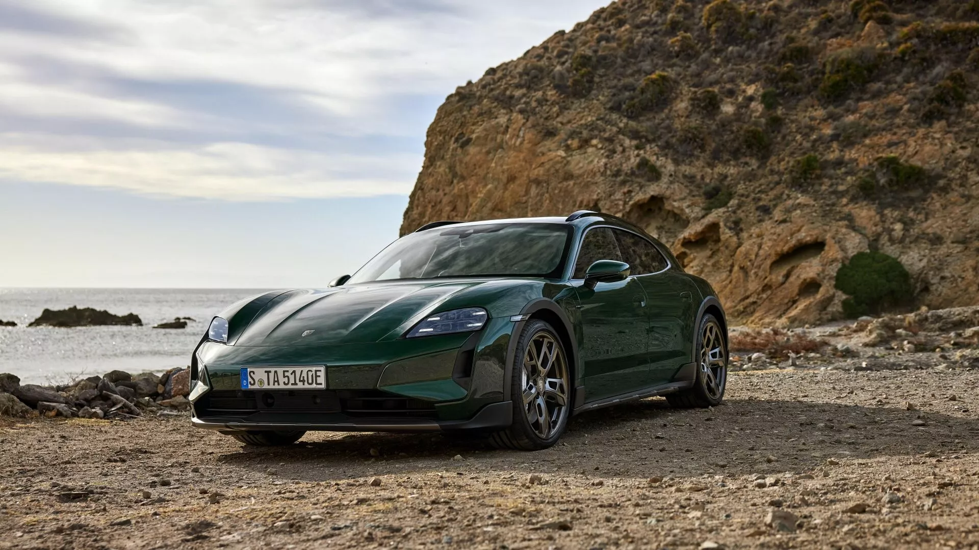 Modèle Porsche Taycan Y1A facelift 2024 photographié en lumière naturelle sur un décor côtier non pavé, présentant sa peinture vert foncé métallique, son design aérodynamique élégant et ses caractéristiques avancées de véhicule électrique contre un fond de roches et de mer.