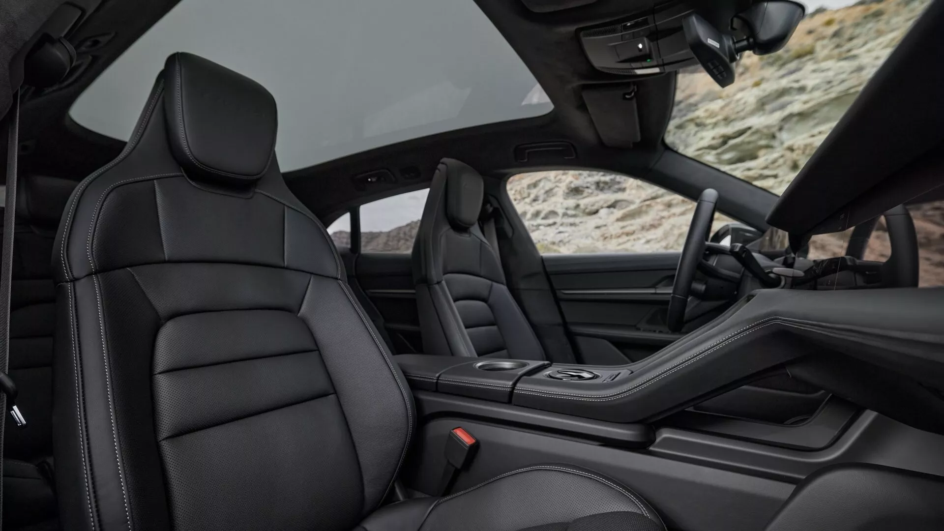 Modèle Porsche Taycan Y1A facelift 2024 montrant la vue intérieure de luxe de la voiture depuis les sièges arrière, se concentrant sur le siège passager avant en cuir noir haut de gamme, console centrale élégante, et indices de conception avancée du tableau de bord et intérieur de porte avec toit convertible rétracté, soulignant l'artisanat, le confort et les caractéristiques technologiques.