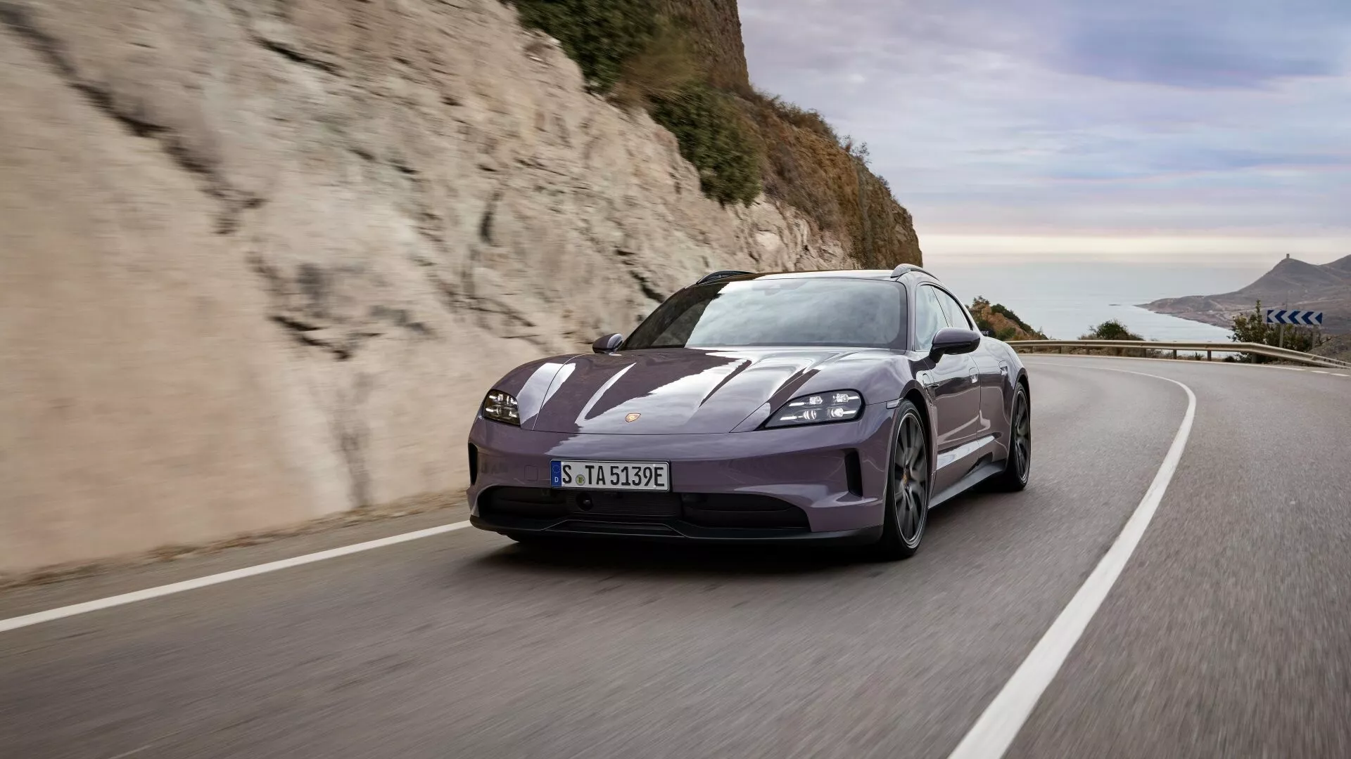 Porsche Taycan Y1A facelift 2024 conduisant sur une route côtière européenne avec vue dynamique avant trois-quarts montrant une posture agressive, un design aérodynamique, des phares LED, des roues en alliage et des pneus de performance sous une lumière du jour éclatante