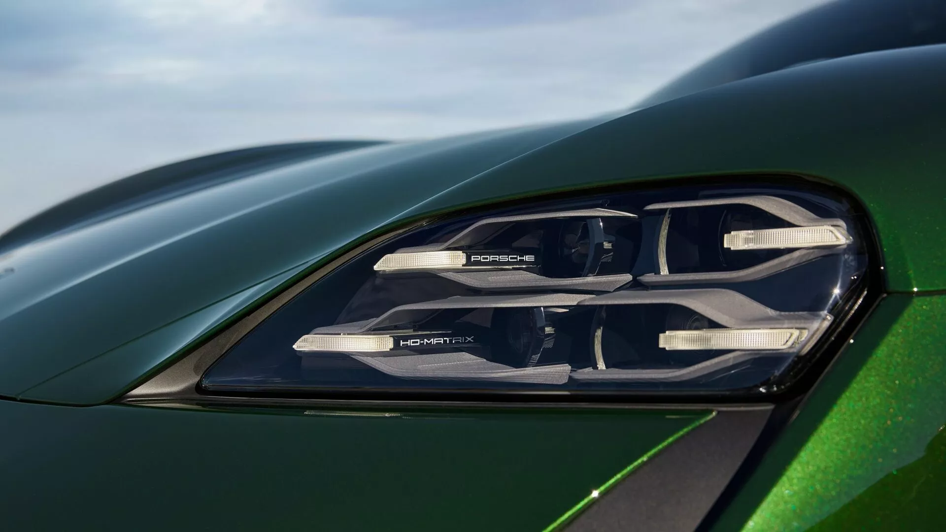 Vue rapprochée du phare de la Porsche Taycan 2024 (restylage Y1A), dévoilant une technologie d'éclairage avancée avec éléments LED, marque PORSCHE et un aperçu du capot et de l'aile en vert métallique reflétant une aérodynamique sophistiquée.