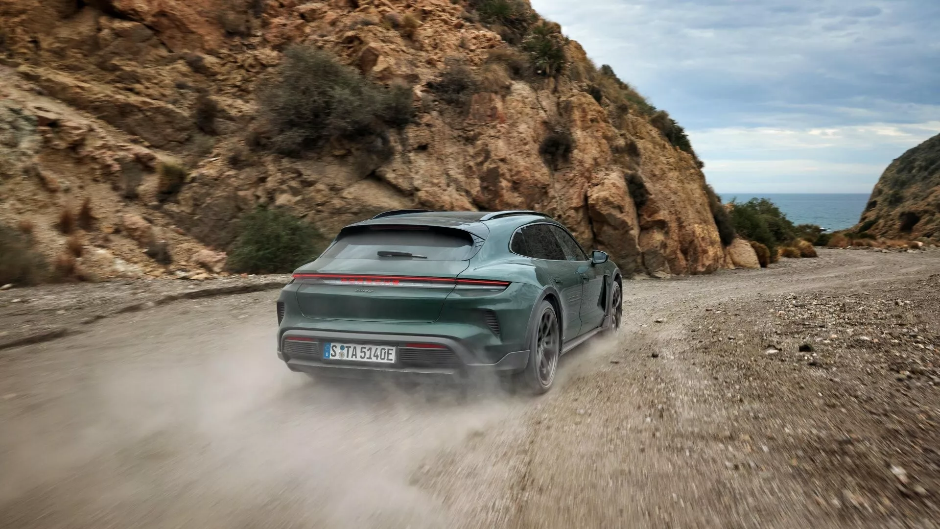 Porsche Taycan 2024 en vert, présentant son design dynamique par une vue arrière trois quarts sur une route de gravier, avec une barre lumineuse arrière distinctive et une carrosserie bicolore, suggérant des capacités de traction intégrale et un groupe motopropulseur électrique, au milieu d'un paysage naturel rocheux.