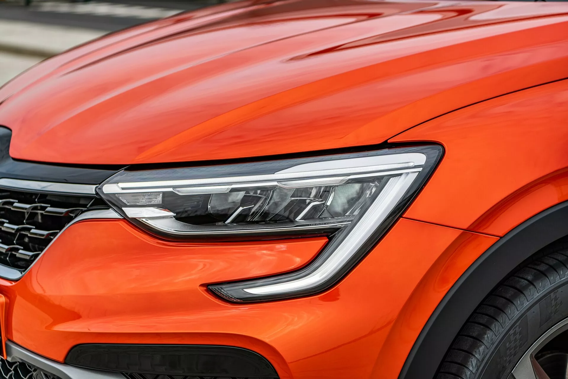 Vue en gros plan de la partie avant du Renault Arkana 2021 orange vif, se concentrant sur les détails de conception modernes, y compris les phares LED avancés, les courbes aérodynamiques du capot, une grille en nid d'abeille partiellement visible et un fragment d'une grande roue en alliage, suggérant des performances haut de gamme et des technologies automobiles de pointe.