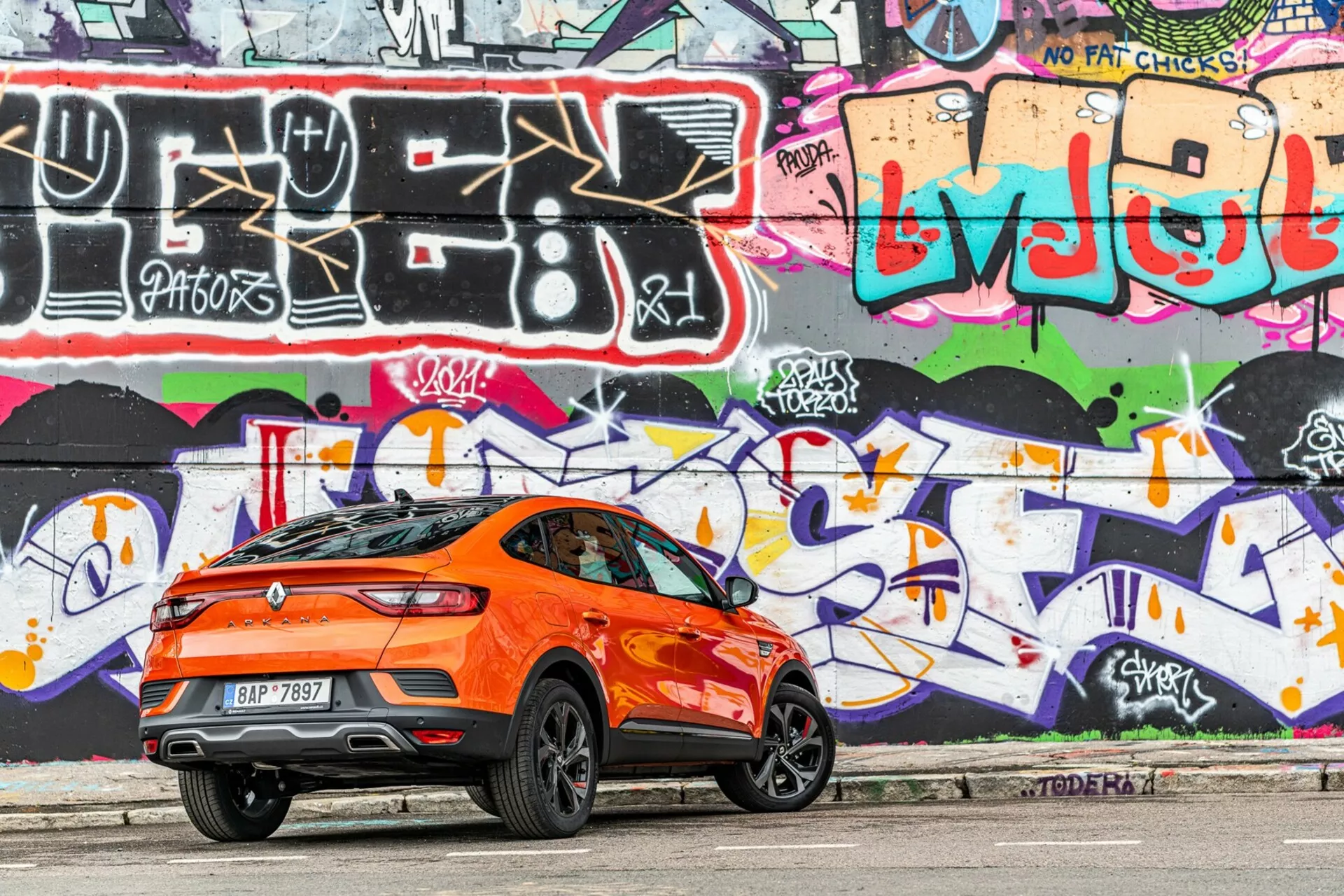 Vue de profil latéral d'un Renault Arkana 2021 de couleur orange vif, garé sur une route urbaine. Photographié depuis le côté droit avec un accent sur les trois quarts arrière, l'image capture la ligne de toit inclinée et sportive de la voiture, le spoiler arrière, la haute ligne de ceinture, les grandes roues en alliage élégantes, les fenêtres teintées et les lignes de caractère aérodynamiques. Les feux arrière sont élégants et probablement à LED. En arrière-plan, on voit un mur vibrant de graffitis, accentuant l'esthétique urbaine. La photo, prise en plein jour, montre des ombres douces sous la voiture, mettant en valeur les détails de son design.