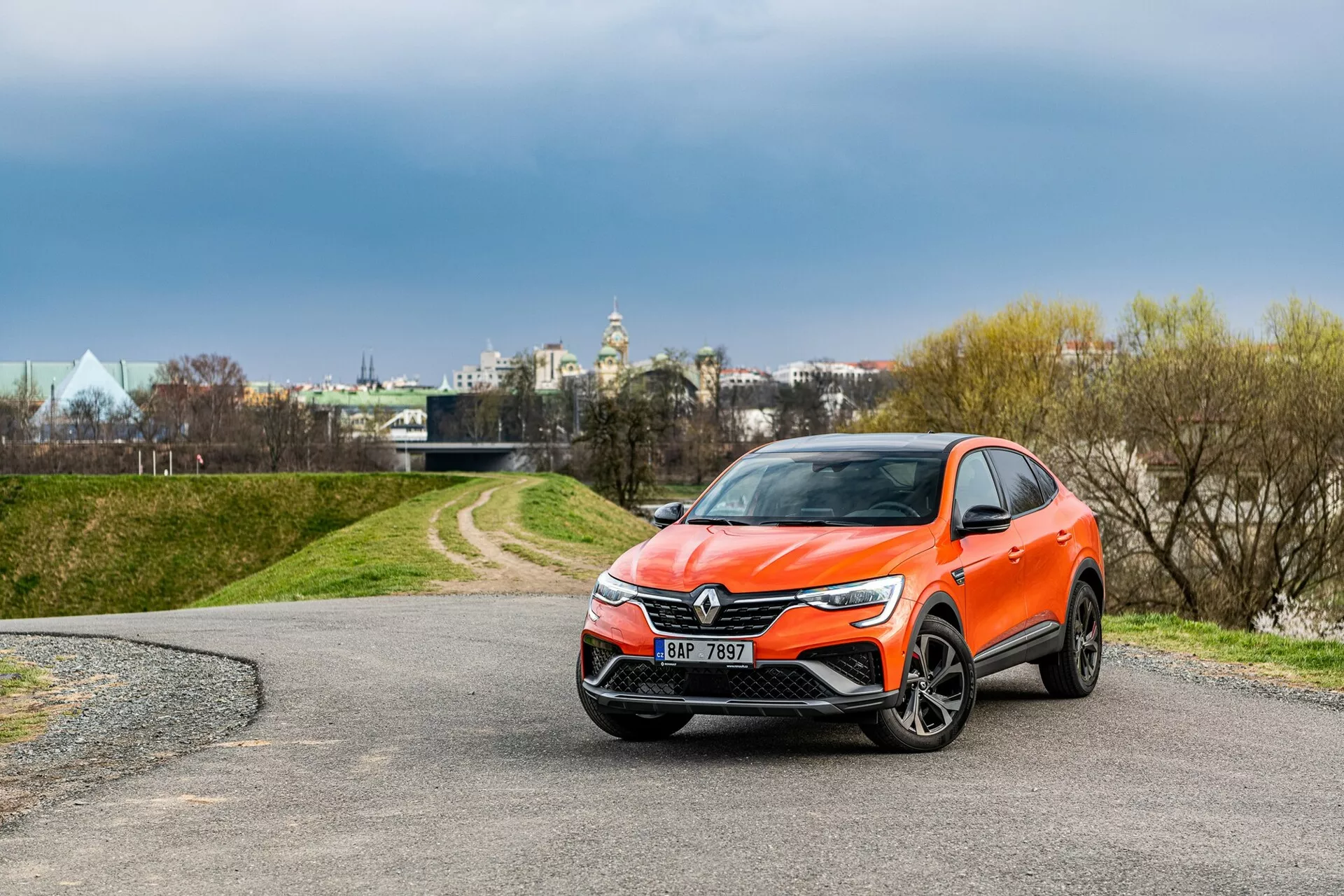 L'image montre un SUV crossover Renault Arkana 2021 moderne, garé sur un chemin sinueux. Capturée d'une vue trois-quarts avant, la photographie affiche clairement la calandre avant avec un emblème proéminent, des phares élégants s'étendant vers les côtés et un pare-chocs avant stylisé probablement équipé de feux antibrouillard et de capteurs. Les rétroviseurs latéraux au design aérodynamique avec des indicateurs de direction intégrés possibles, les roues en alliage et une carrosserie orange vibrante rehaussent l'apparence générale. L'environnement comprend des arbres et un ciel nuageux en arrière-plan, avec des structures suggérant une zone urbaine au loin.